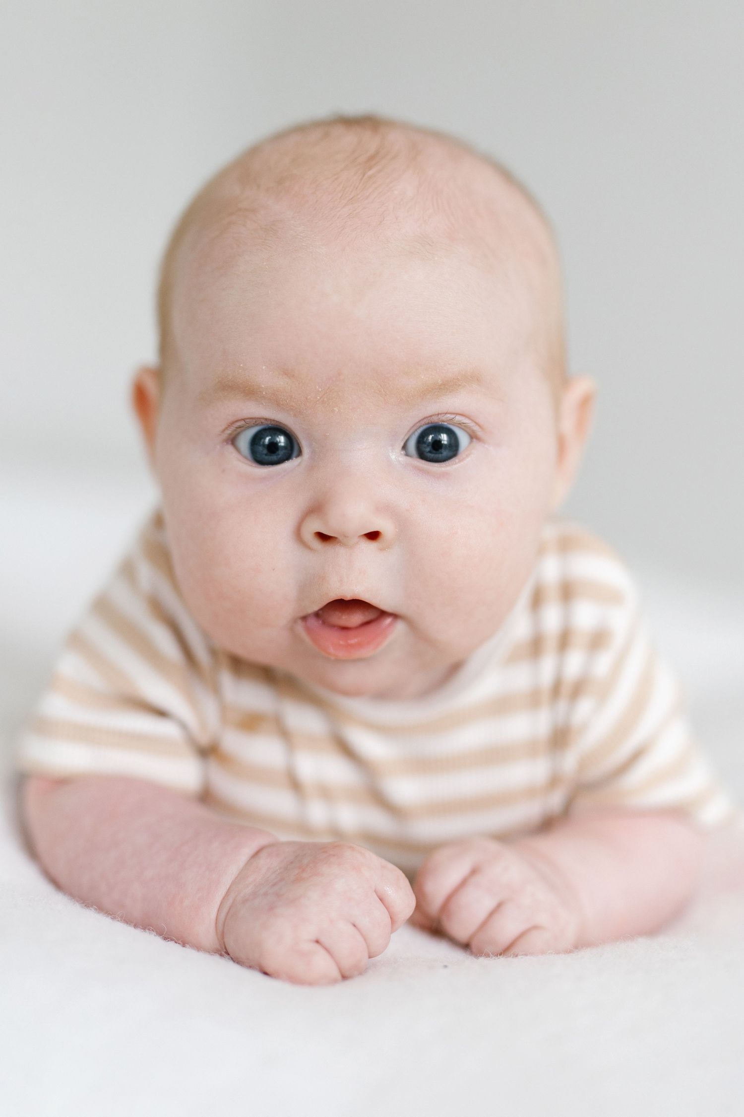 Adorable baby with blue eyes lying on tummy wearing a beige striped onesie, looking curiously at the camera.