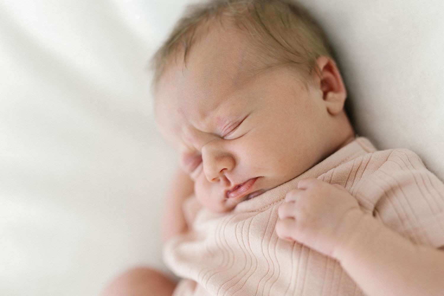 Peaceful newborn baby sleeping in a soft pink onesie on a white blanket, with tiny hands tucked under chin.