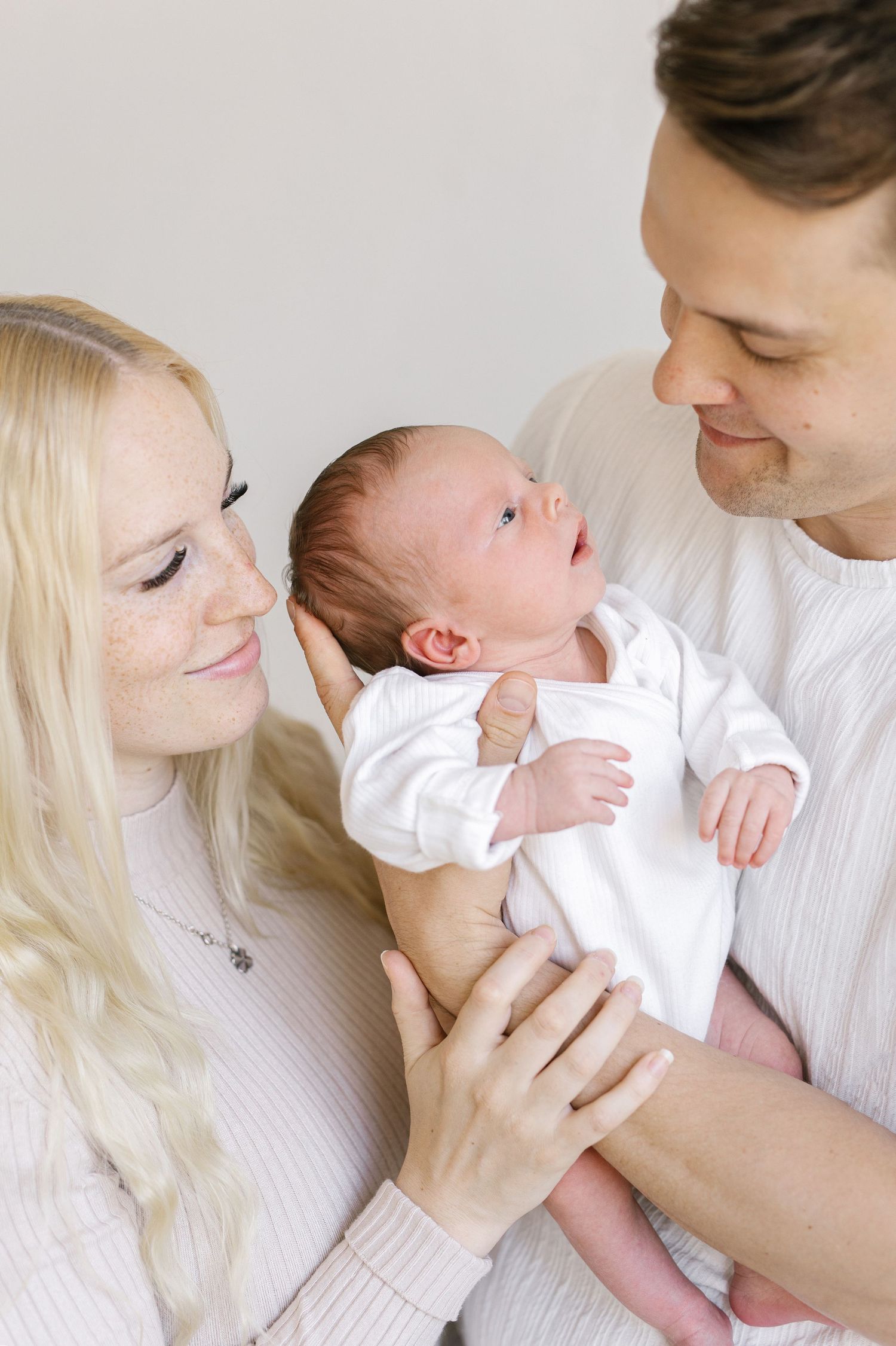 New parents lovingly hold their newborn baby in white outfits against a light background.