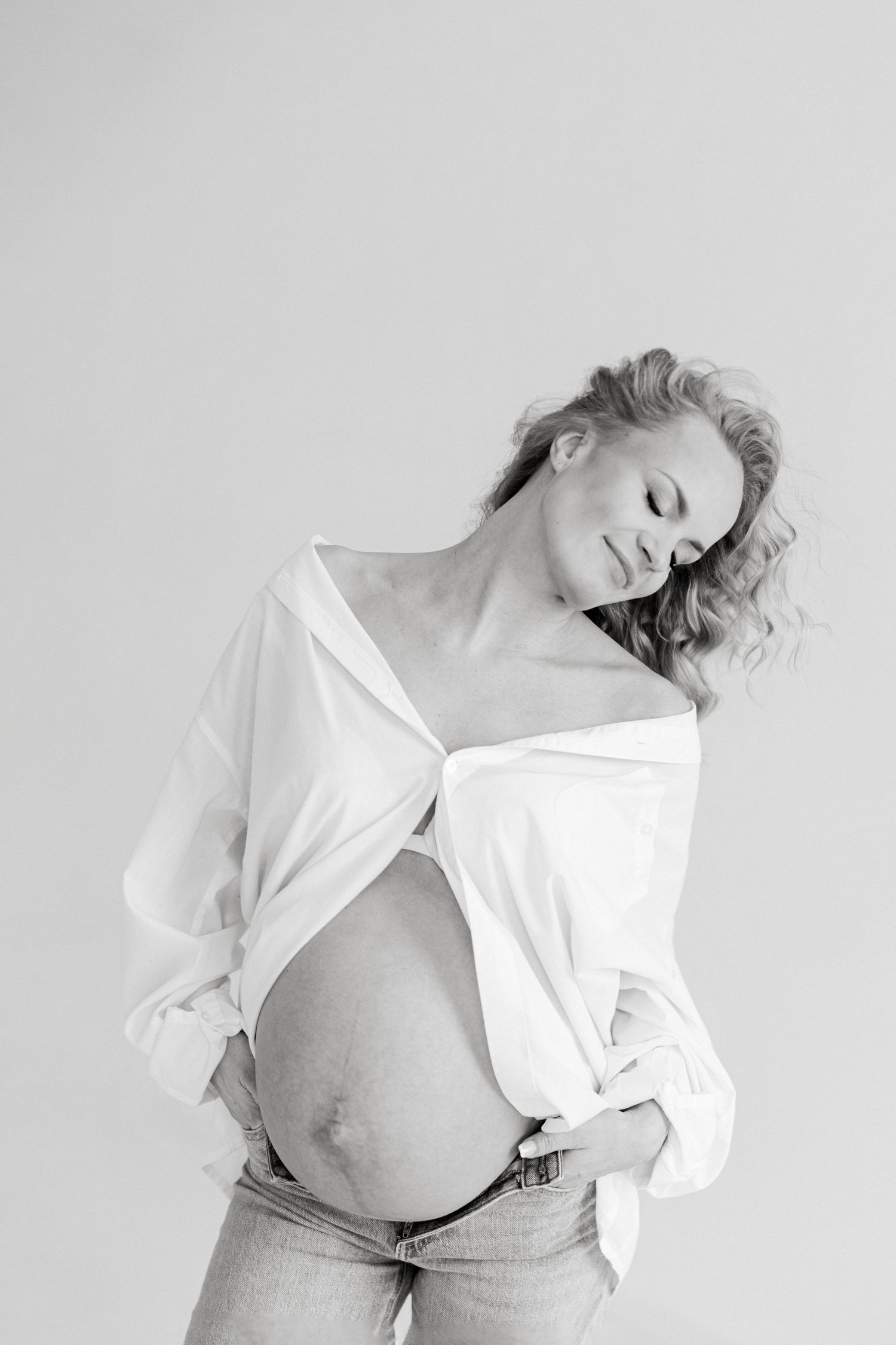 Black and white portrait of a pregnant woman in an open white shirt and jeans, cradling her baby bump.