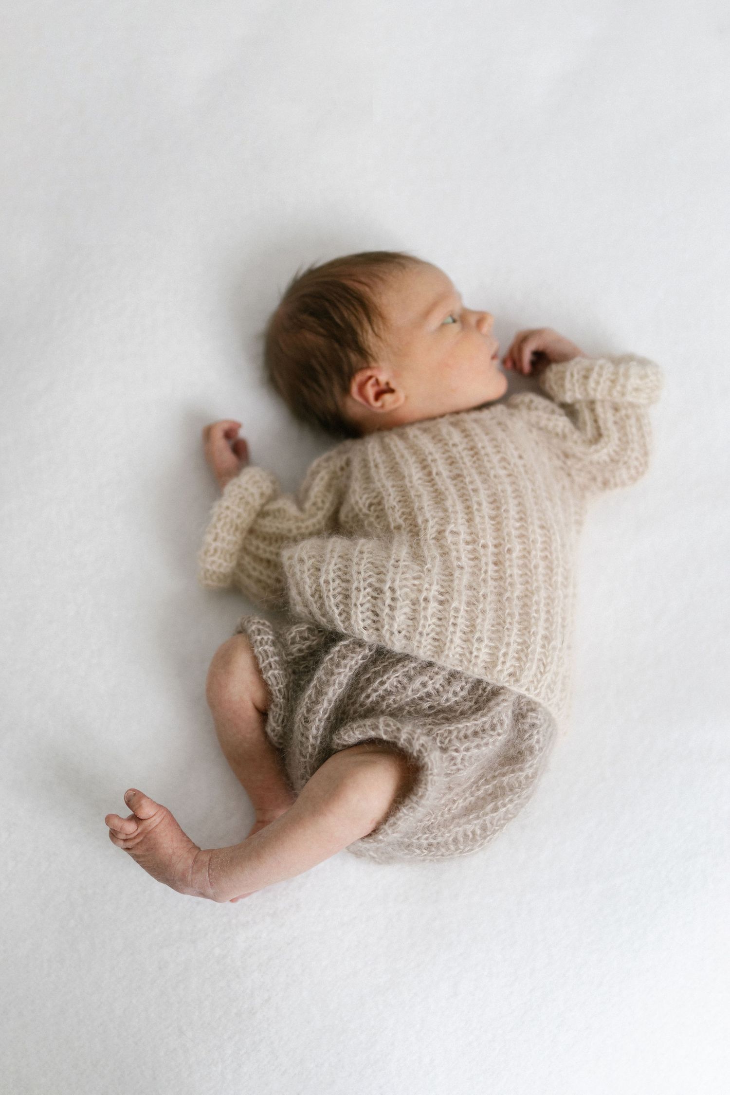 Sleeping newborn baby in a beige knit romper lying on a white blanket, photographed from above.