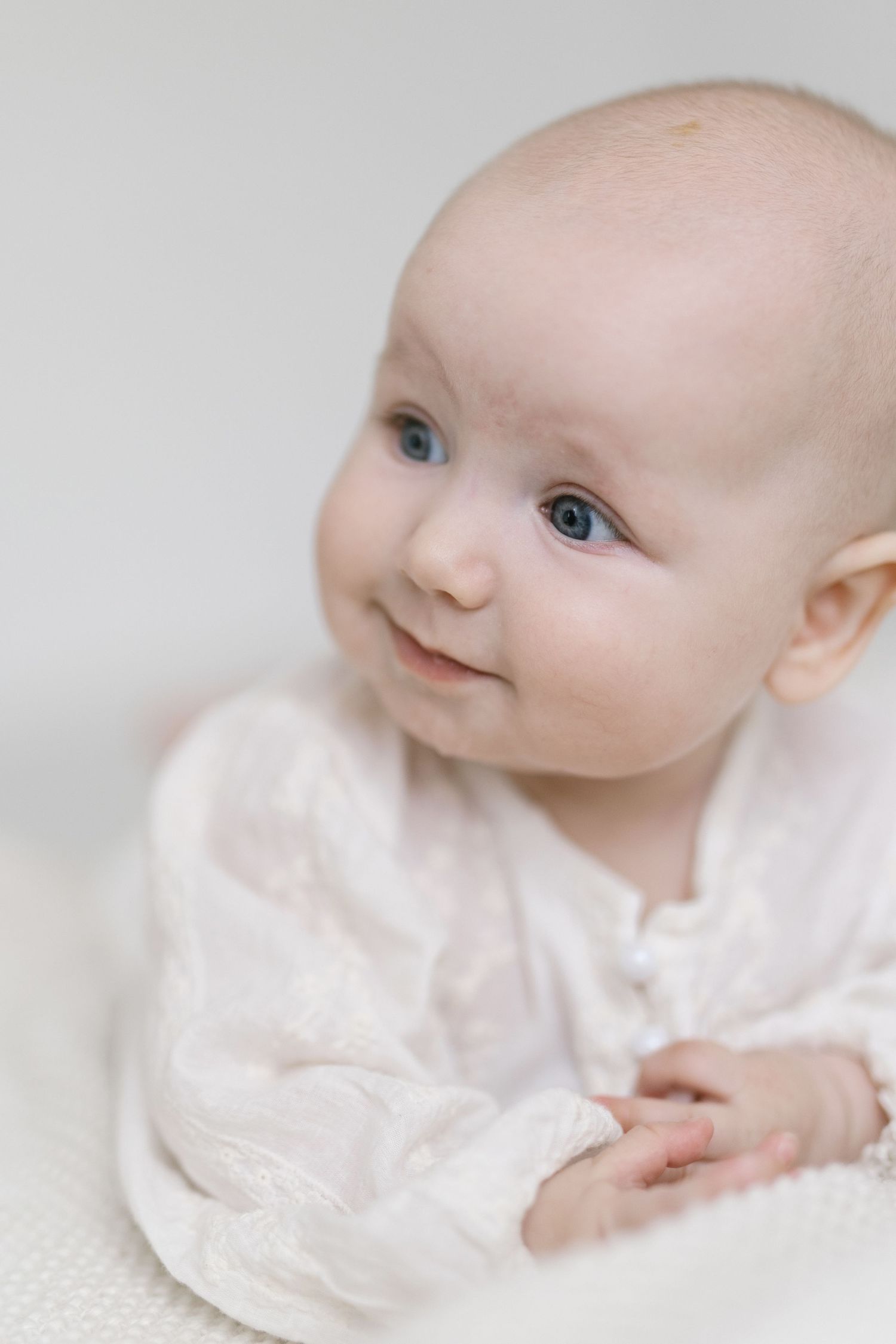 Adorable baby with blue eyes wearing a white lace outfit, smiling softly against a light background.