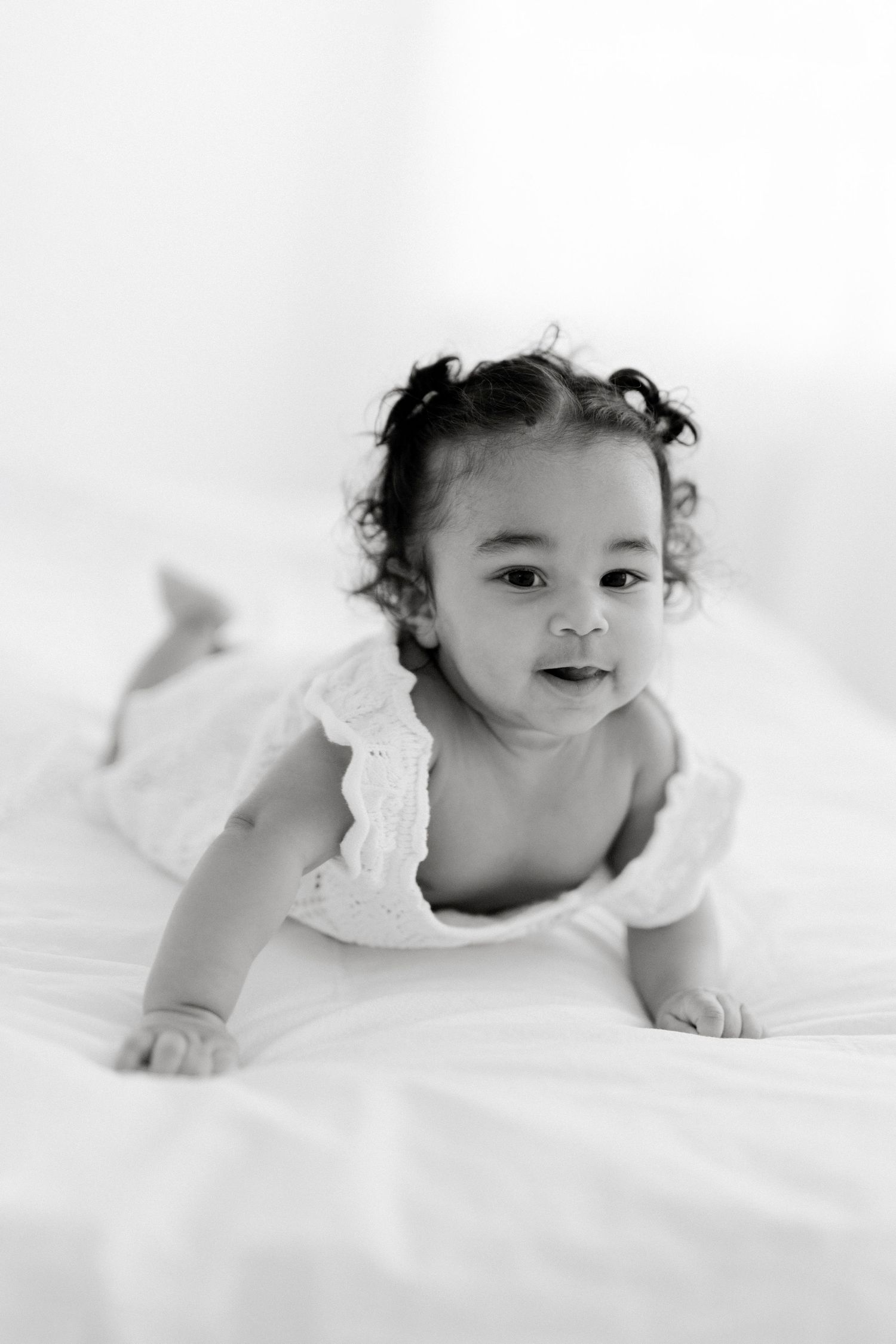Black and white photo of a cute baby girl in a white dress doing tummy time on a white bed.