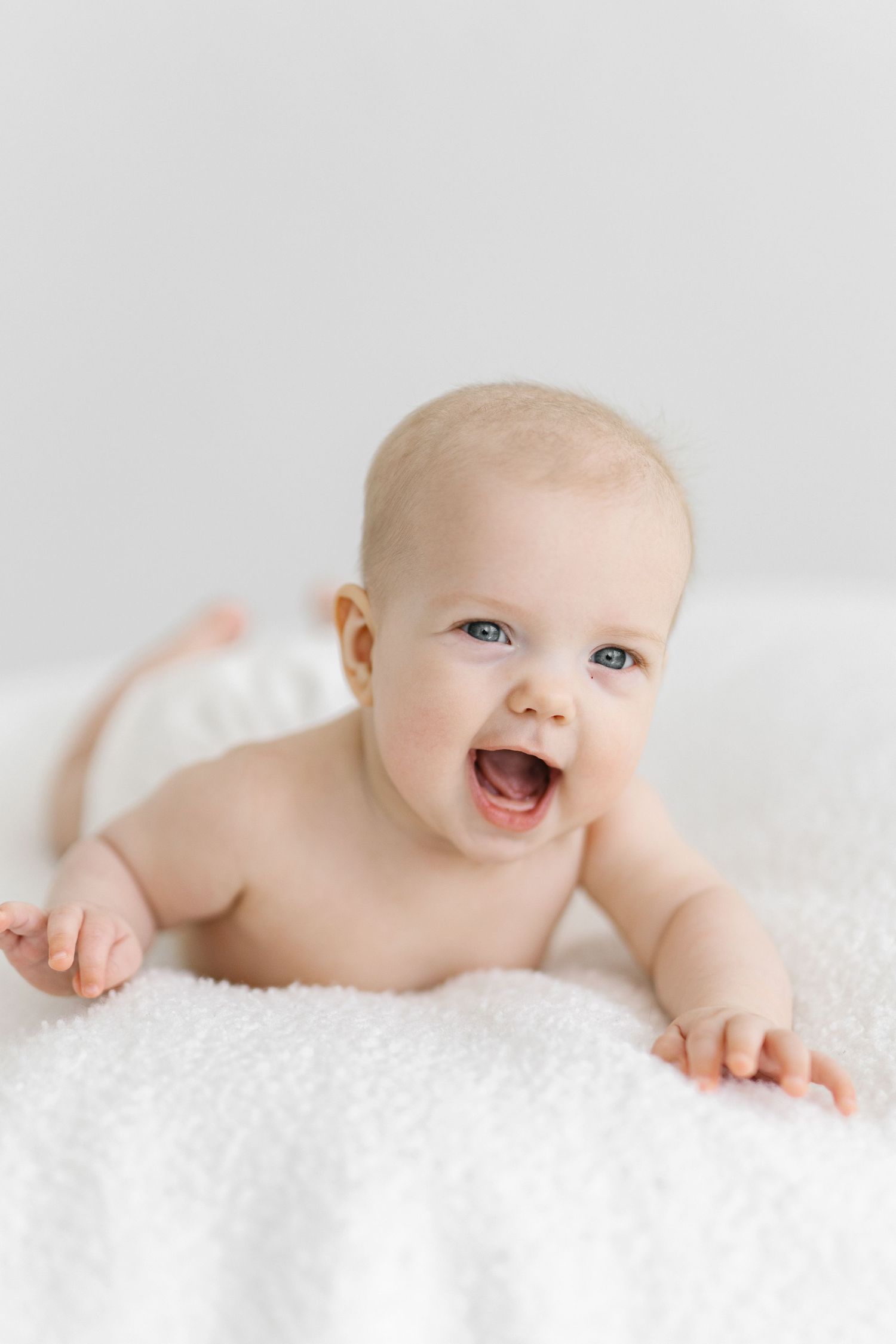 Happy baby lying on white fluffy blanket, laughing with bright blue eyes, wearing a diaper in a bright white studio.