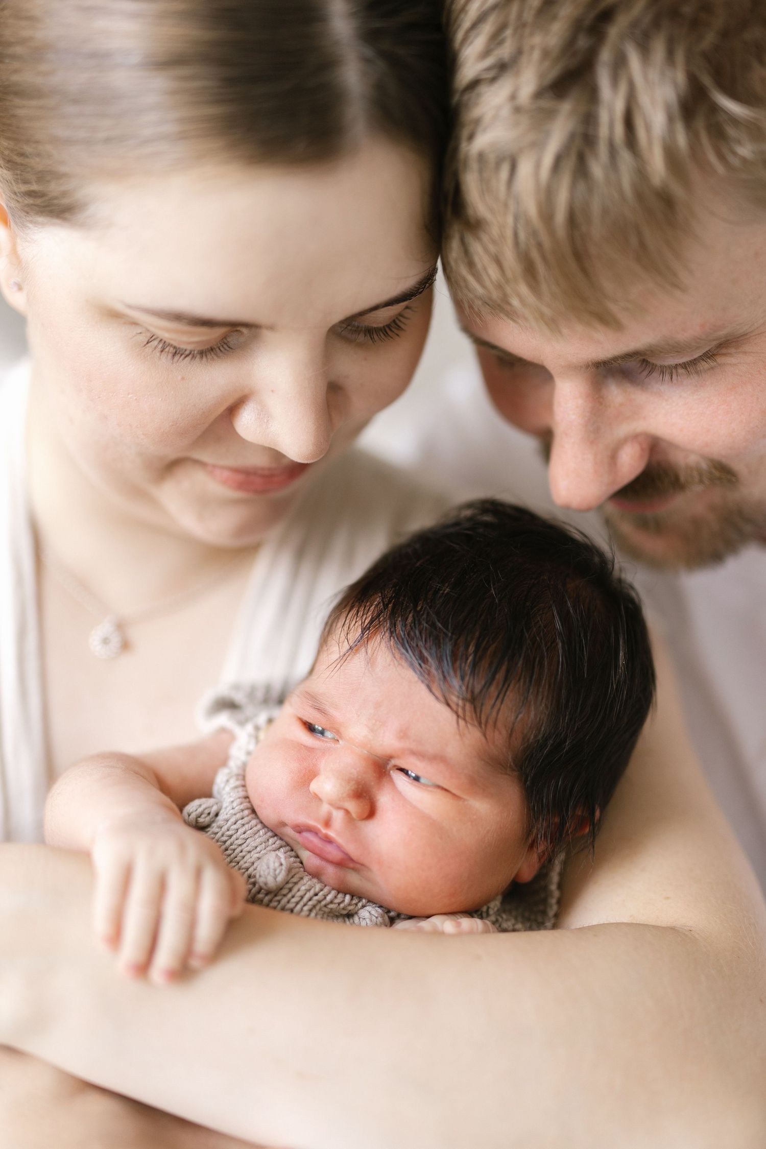 New parents lovingly gaze at their sleeping dark-haired newborn baby wrapped in a knit outfit.