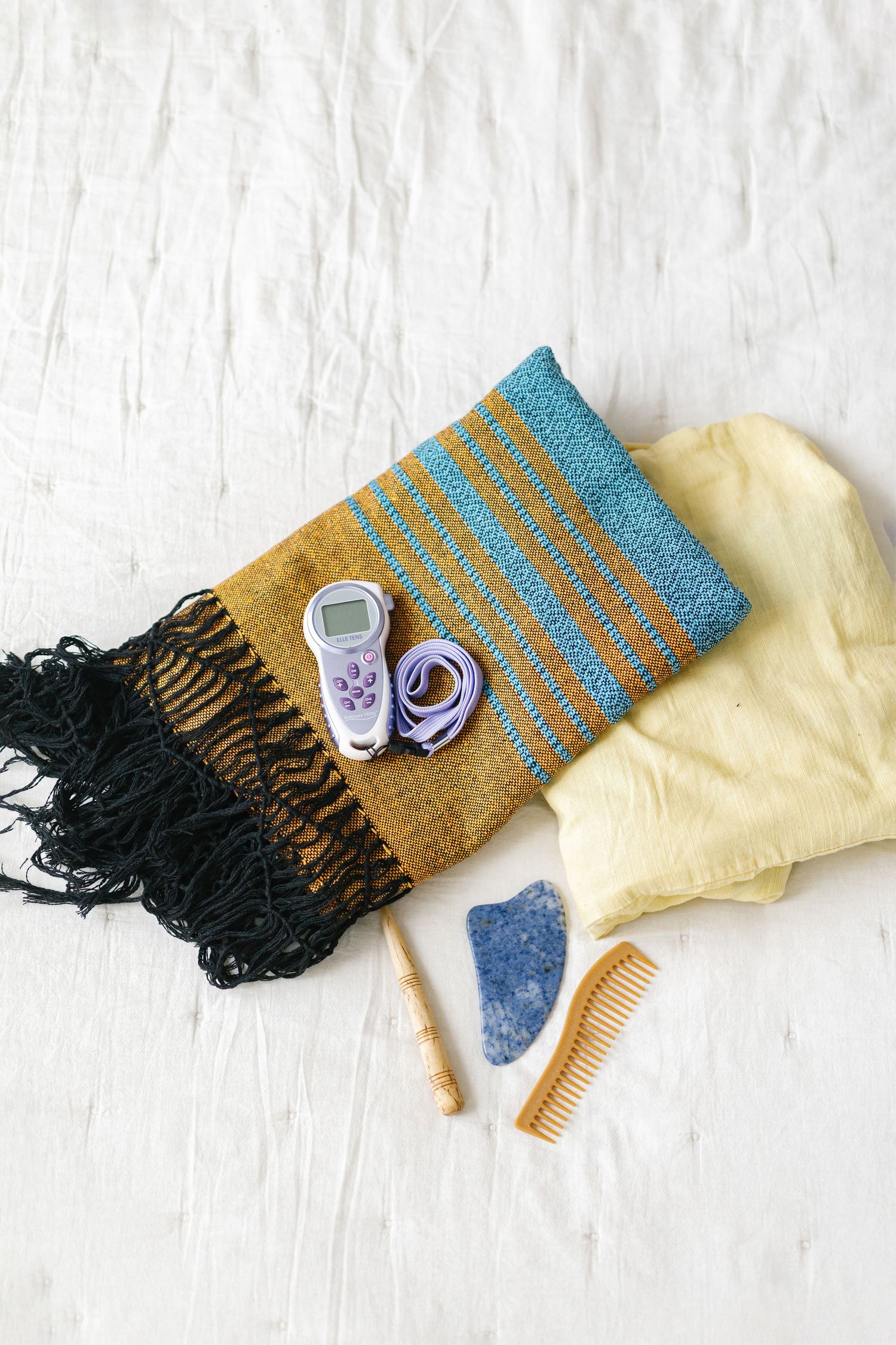 Wellness accessories flat lay featuring a woven scarf, gua sha stone, wooden comb, pulse oximeter, and yellow cloth on white.
