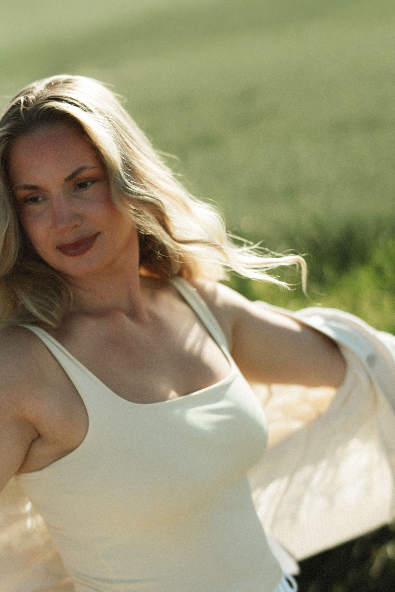 Blonde woman in white tank top smiling outdoors in sunlit green field, hair blowing in breeze.