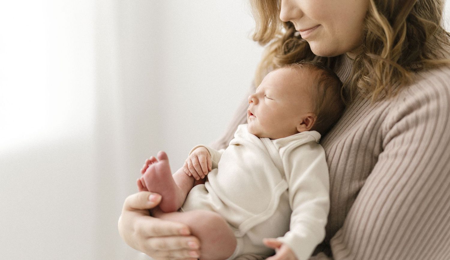 A mother tenderly holds her sleeping newborn baby against her chest near a bright window.