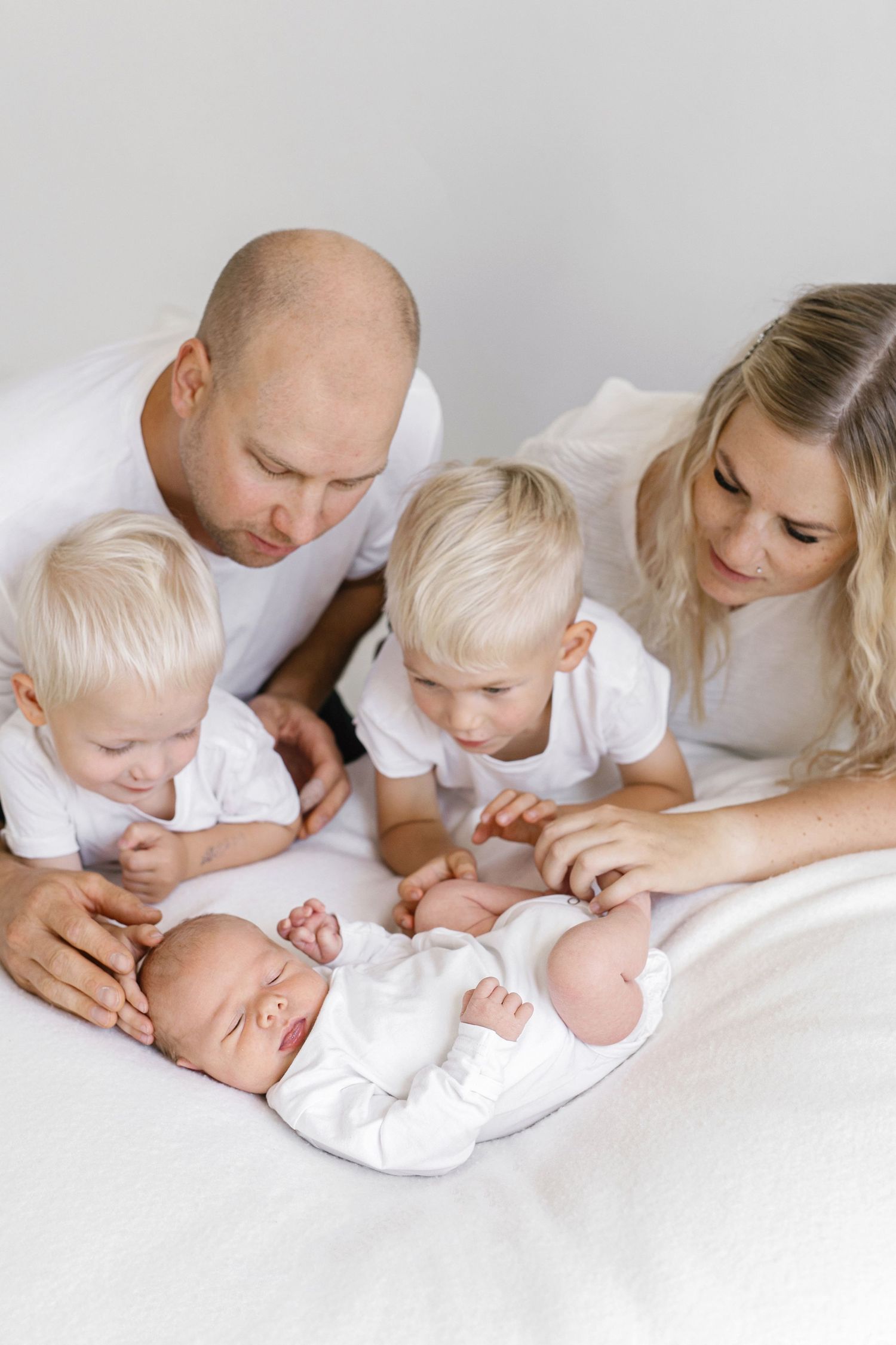 A family of five in white clothing gazes lovingly at their newborn baby lying on a white bed.
