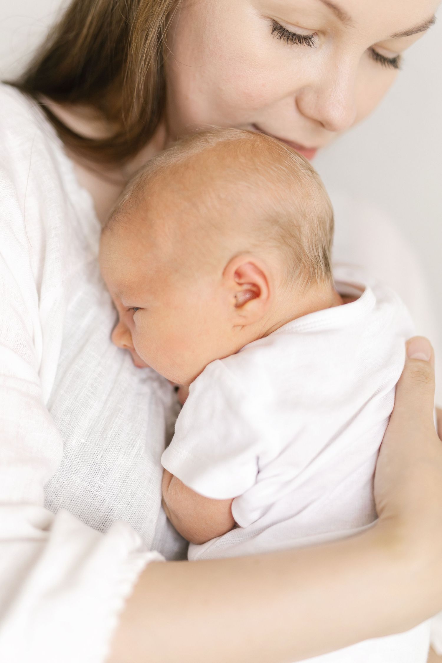 A tender moment as a young mother in white gently cradles her sleeping newborn baby against her chest.