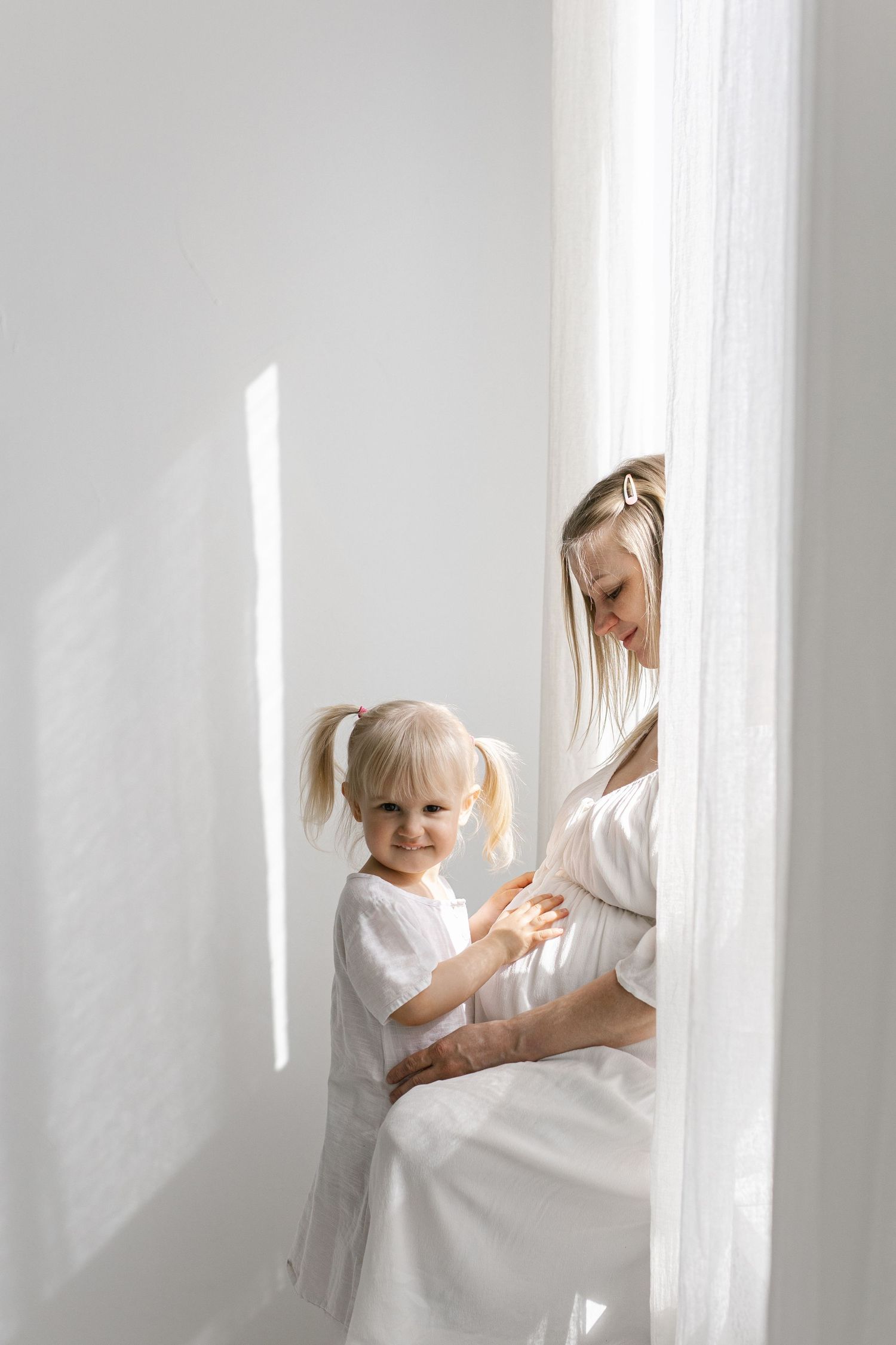 Pregnant woman in white dress with blonde toddler girl by sheer curtains in bright white room.