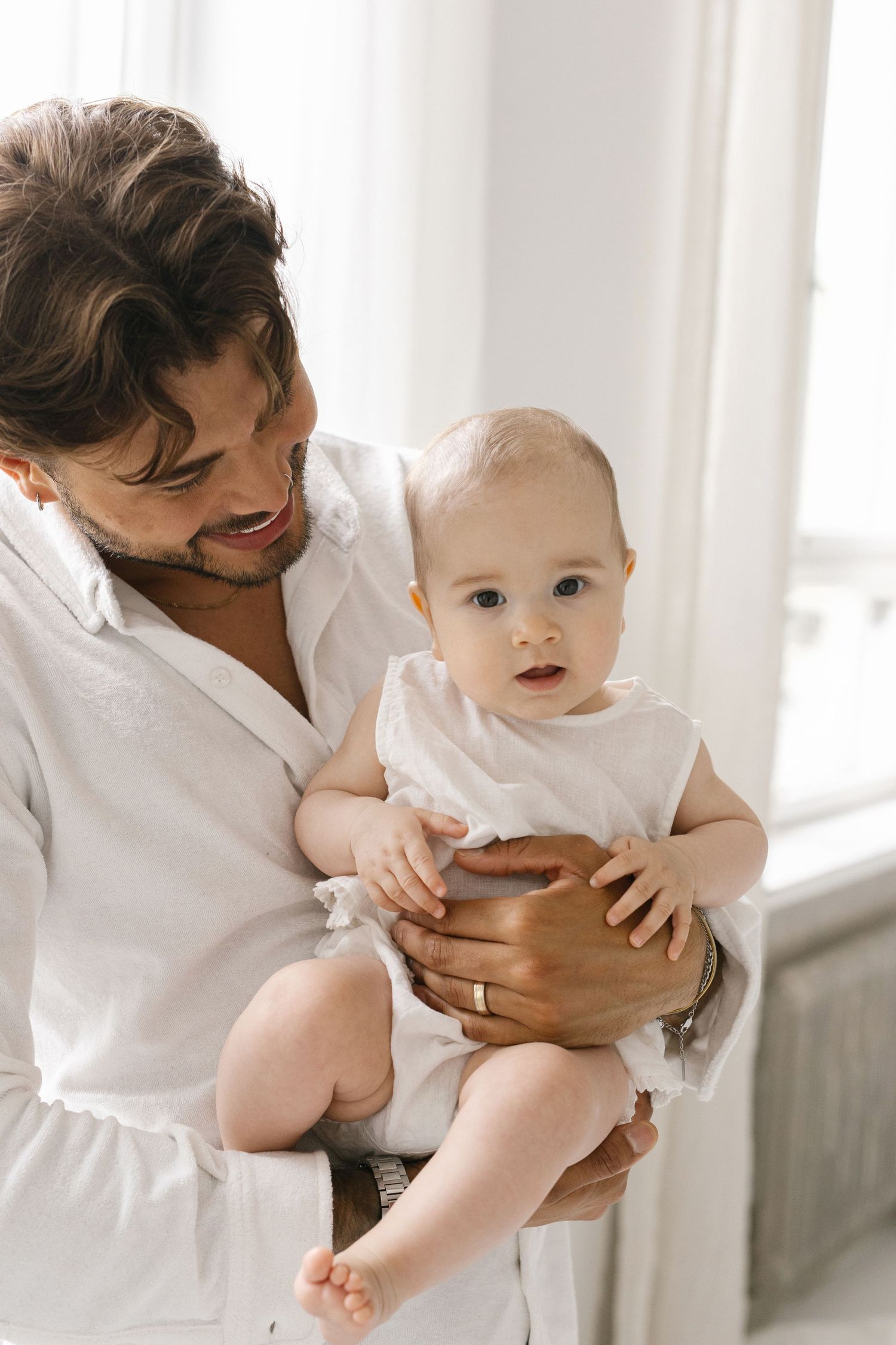 A smiling father in a white shirt tenderly holds his baby in a white onesie near a bright window.