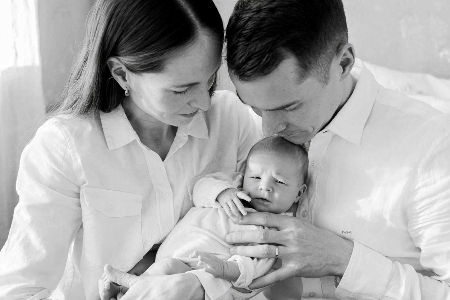 Black and white photo of new parents in white shirts tenderly holding and kissing their newborn baby.