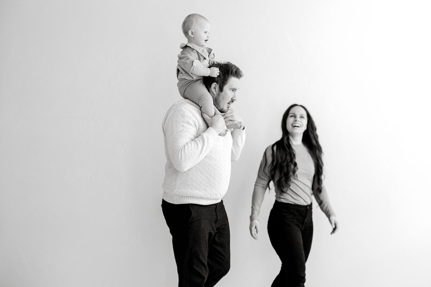 Black and white family photo of parents and baby, father carrying infant on shoulders while mother laughs joyfully.