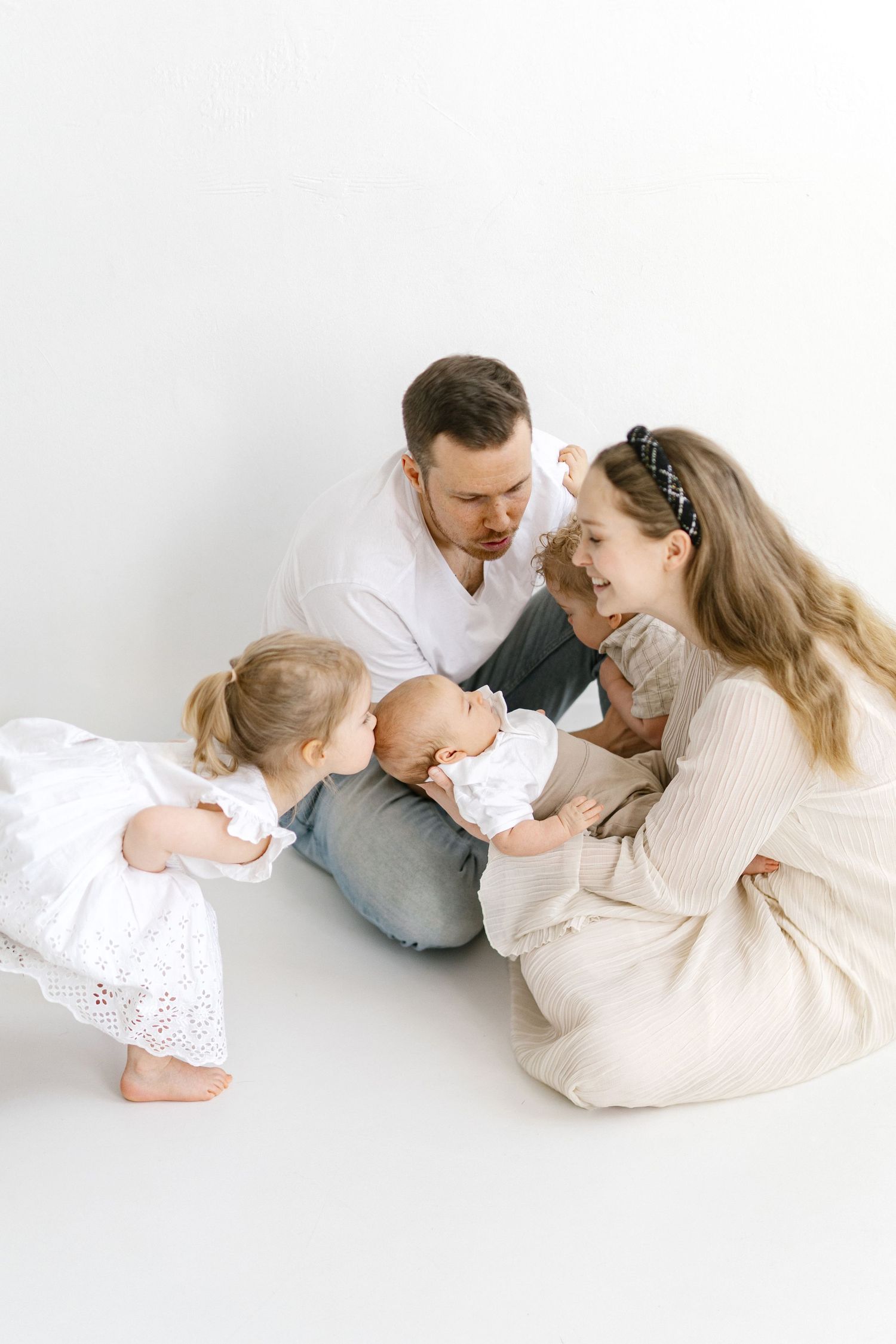 A happy family of four sits on a white studio floor, parents admiring their newborn baby while a toddler girl looks on.