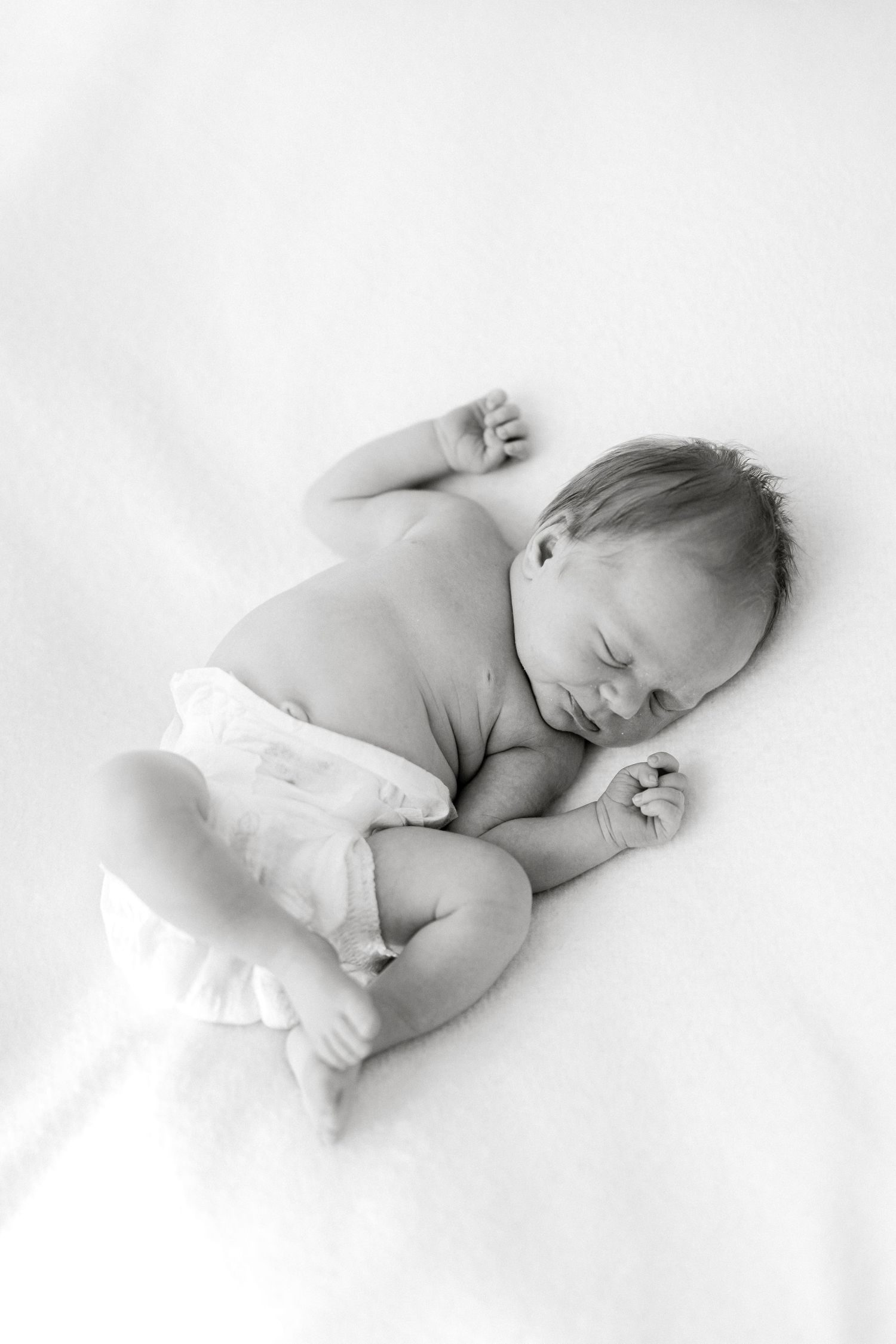 Black and white photo of a peaceful newborn baby sleeping on a white blanket in a natural curl position.