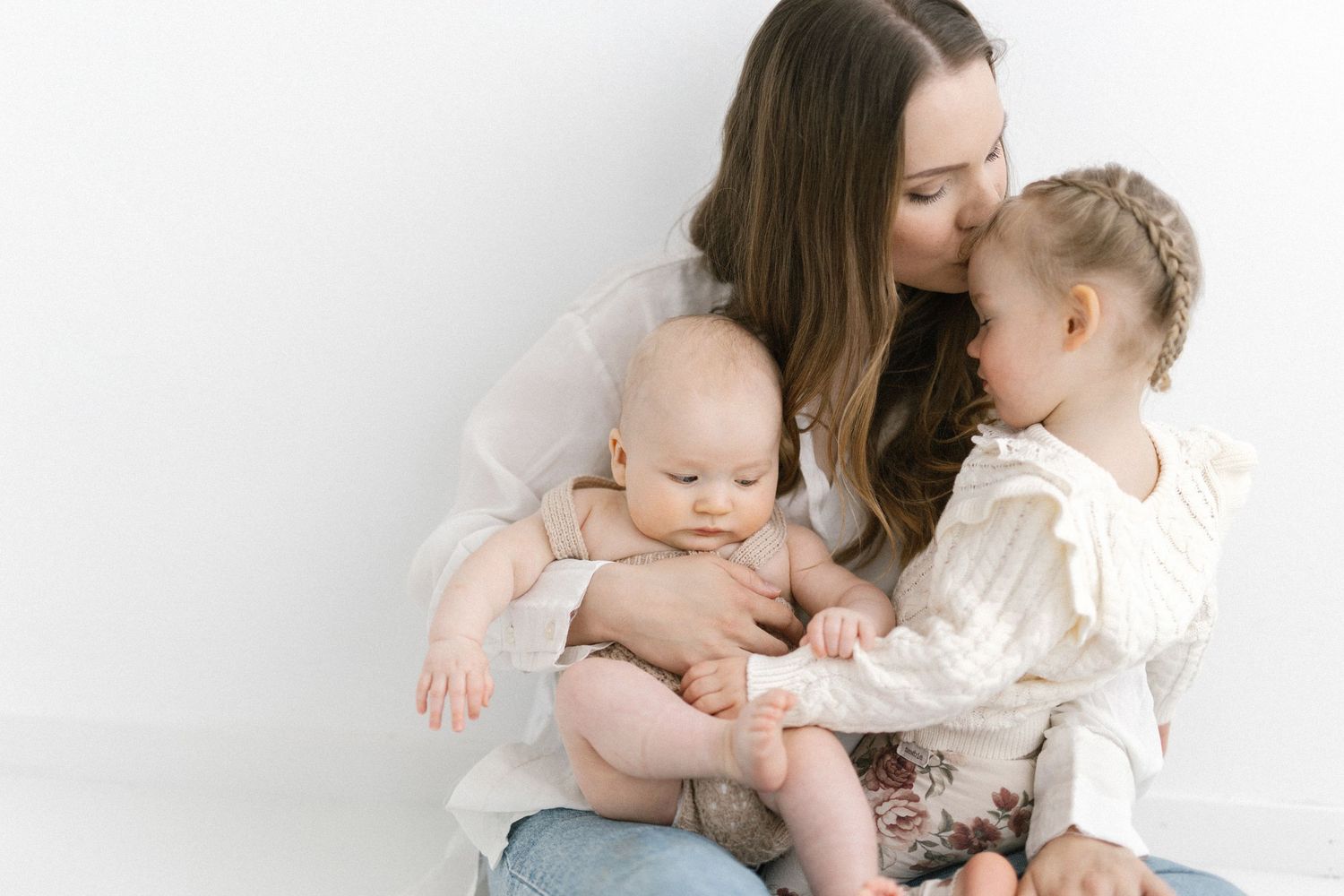 Mother in white blouse kisses toddler girl while holding baby on lap against white background.