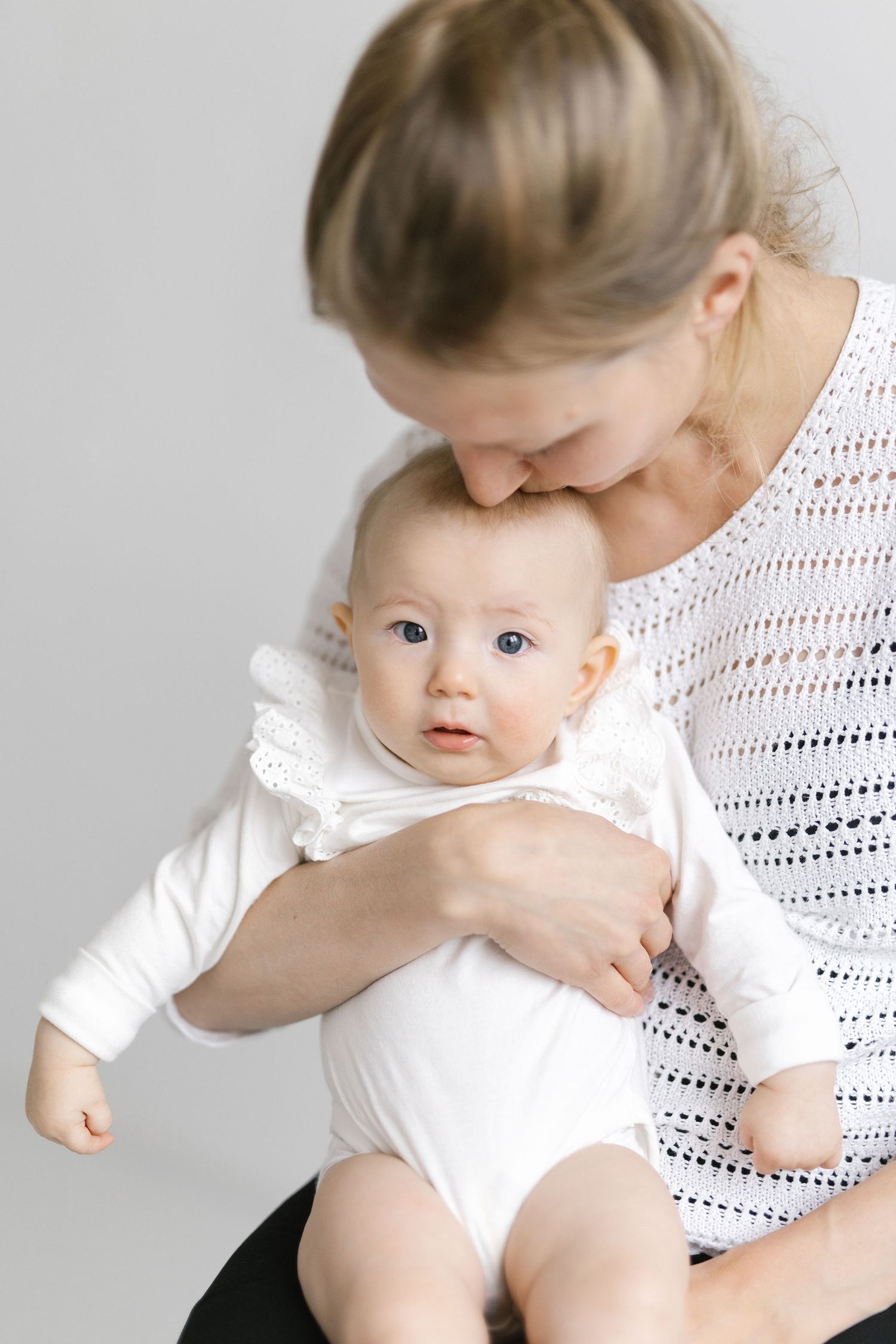 A tender moment as a blonde mother in white holds her wide-eyed baby dressed in a white onesie against a light background.