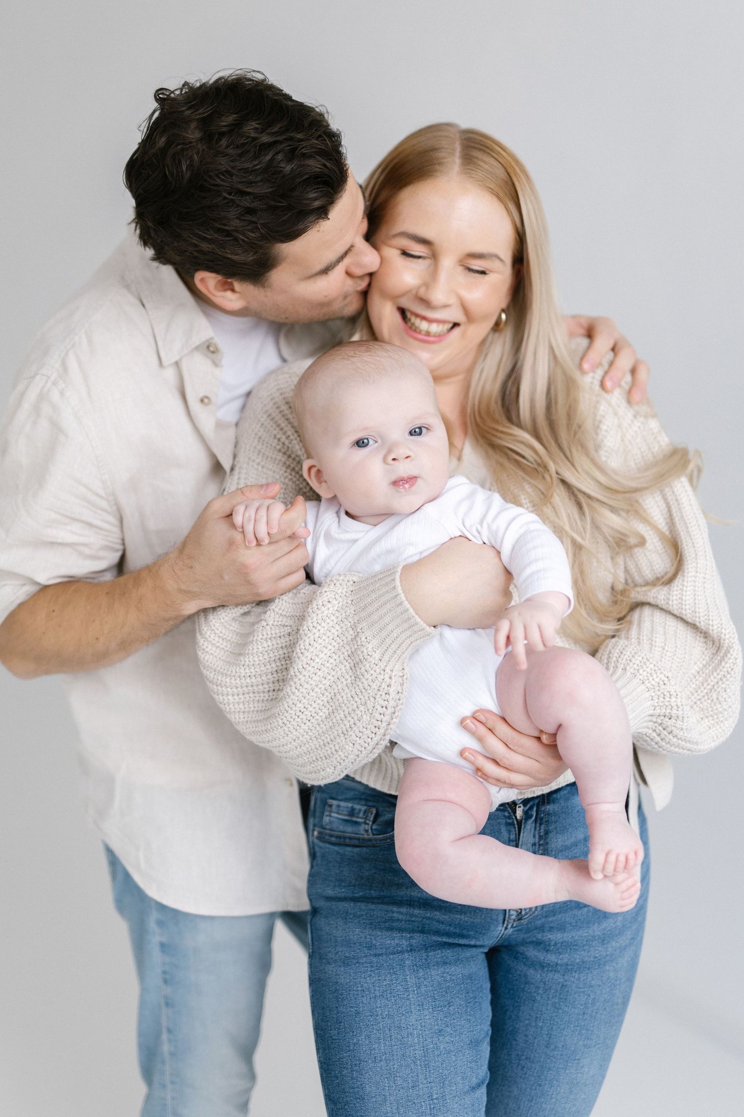 Happy young family with man kissing smiling blonde woman holding adorable blue-eyed baby in white studio setting.