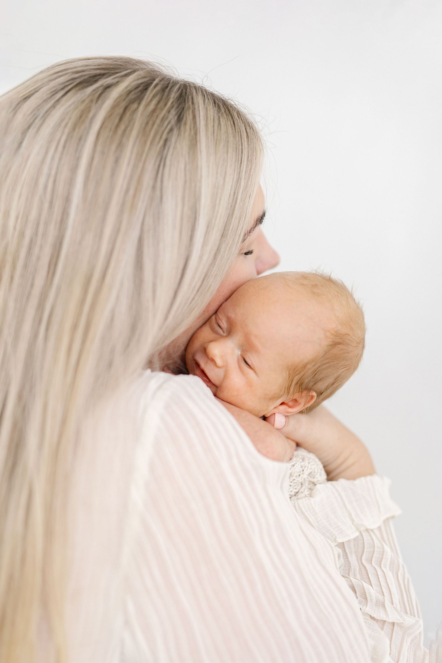 A mother with blonde hair tenderly kisses her sleeping newborn baby, both dressed in soft white clothing.