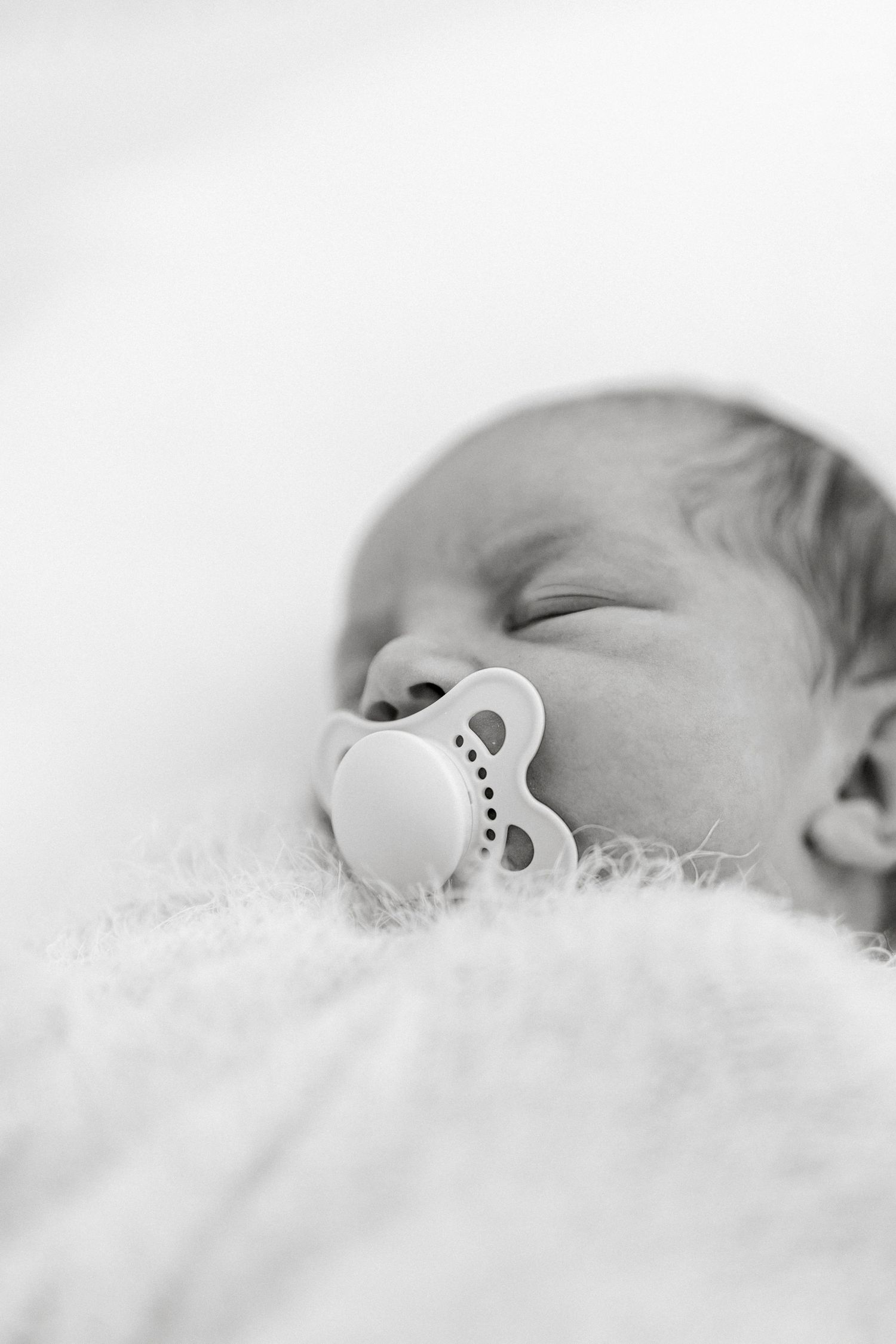 Black and white photo of a sleeping newborn baby with a pacifier, lying on a soft white blanket.