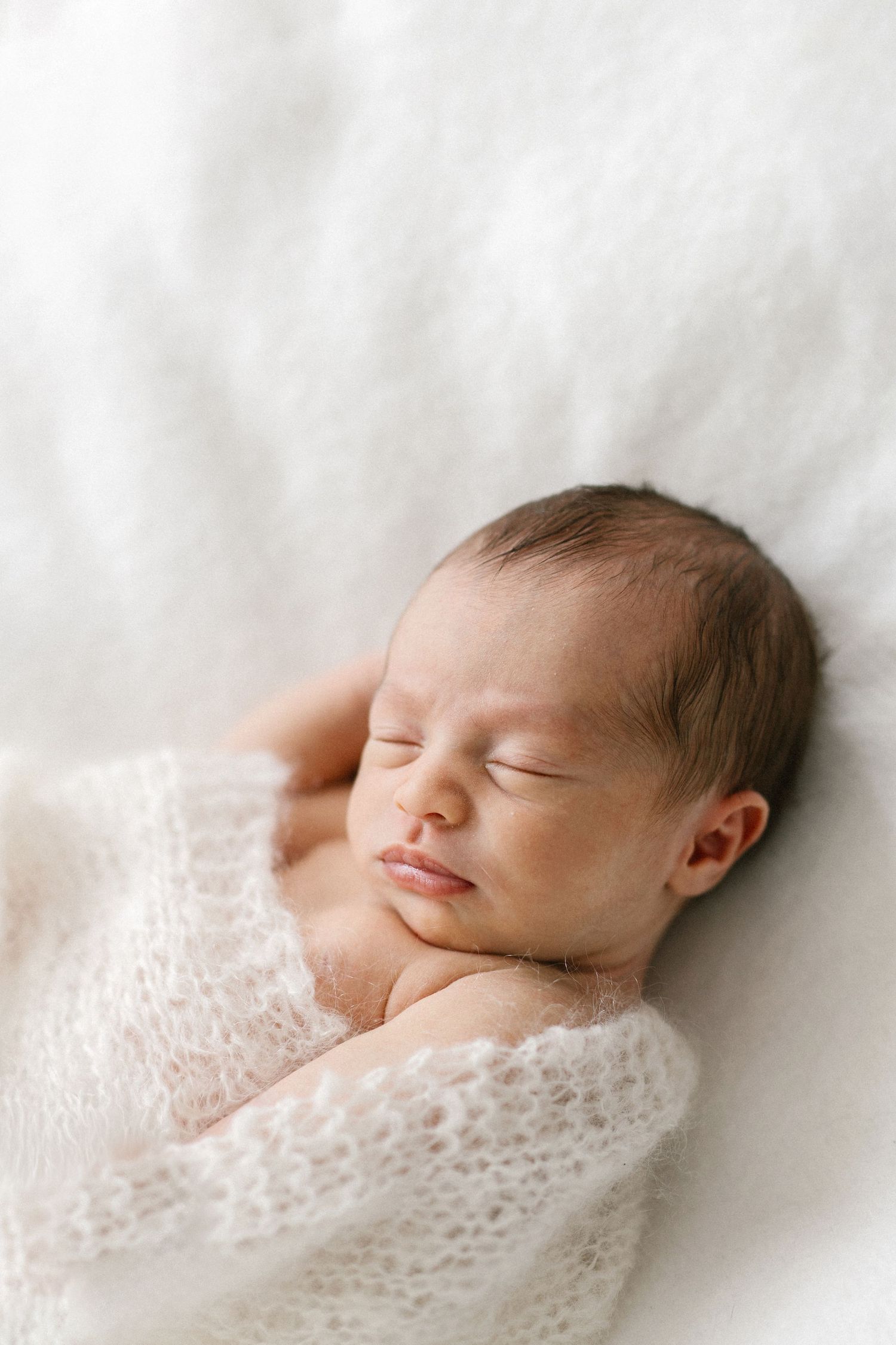Peaceful newborn baby sleeping wrapped in a soft white knit blanket on a fluffy white background.