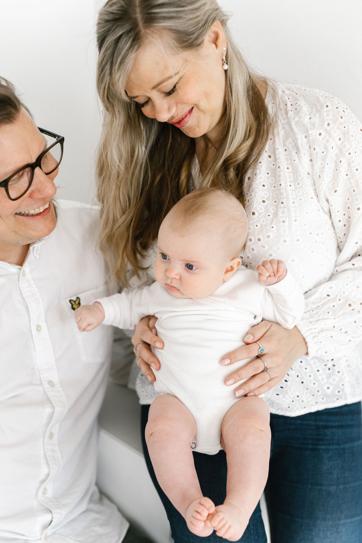 Two smiling parents in white tops lovingly hold their alert baby dressed in a white onesie.