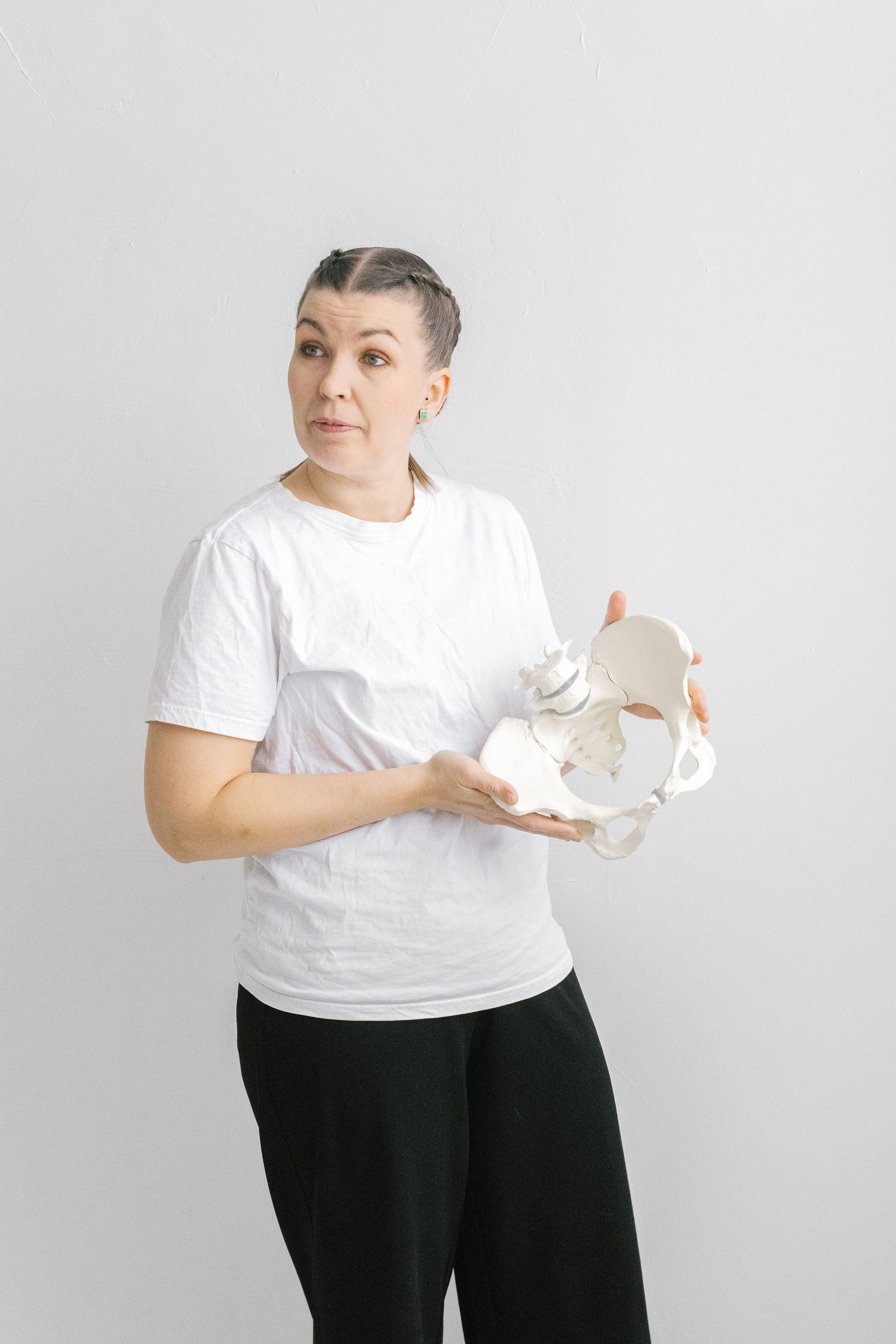 Woman in white t-shirt holding a human pelvis anatomical model against a light gray background.