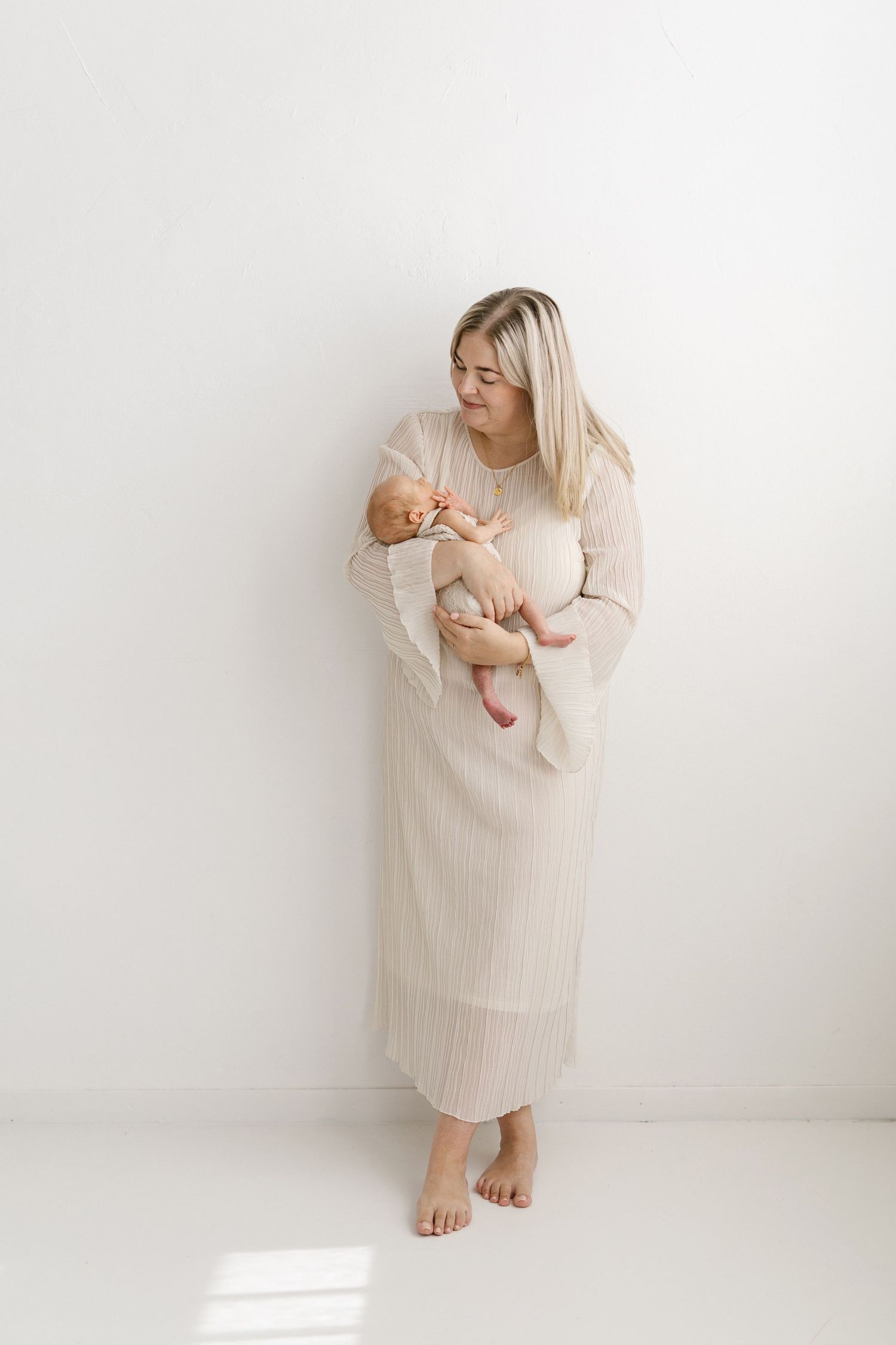 Barefoot blonde mother in beige ribbed maxi dress tenderly holds sleeping newborn baby against white studio wall.