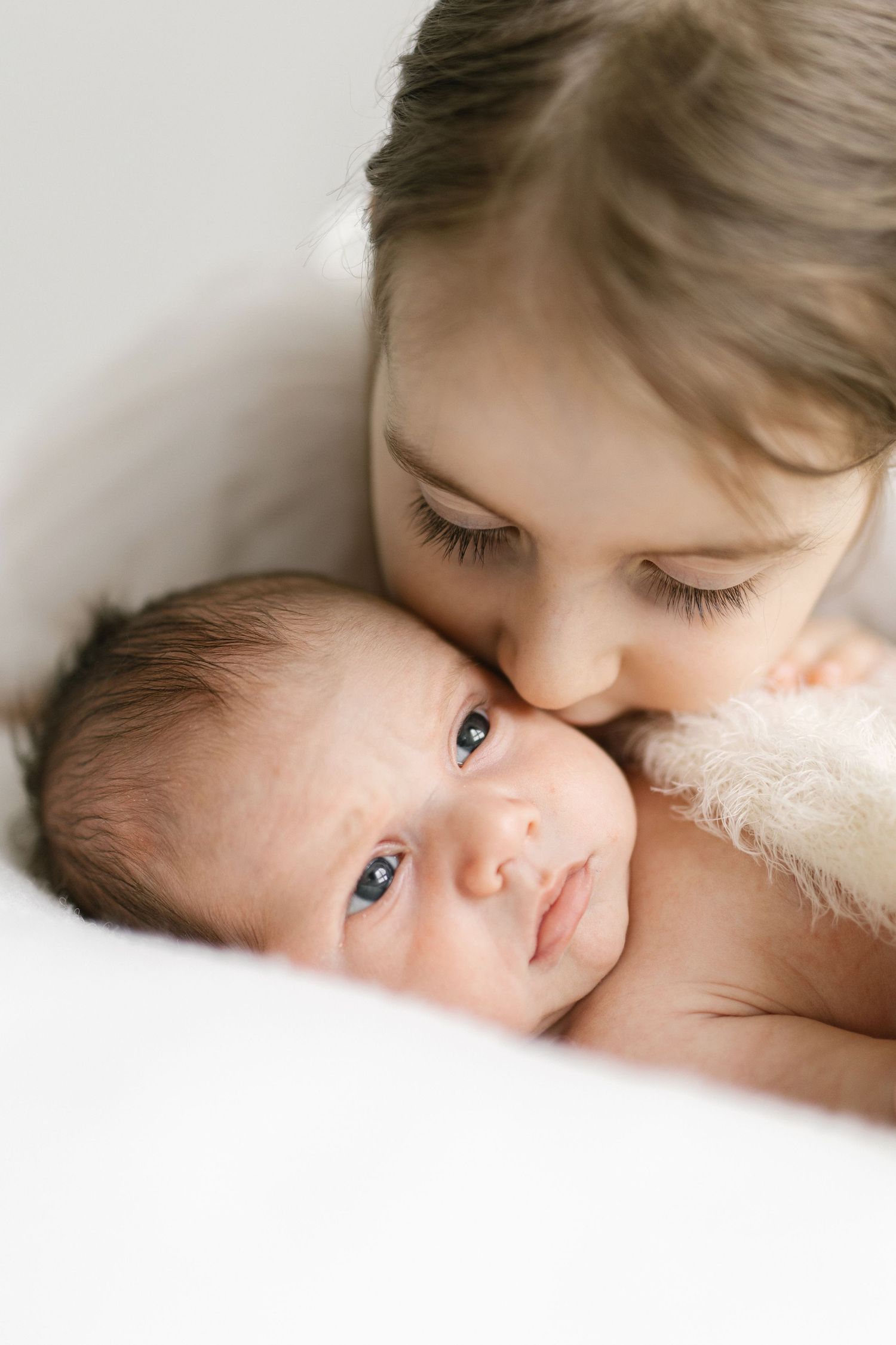 Older child gently kissing a newborn baby on the head while the infant looks at the camera.