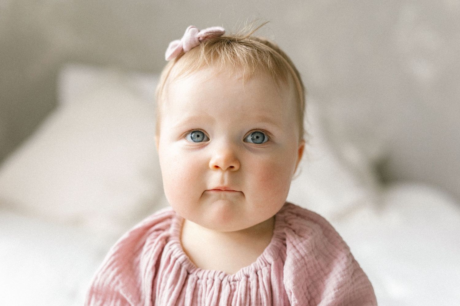 Adorable baby girl with blue eyes wearing a pink dress and bow headband, sitting against a white background.