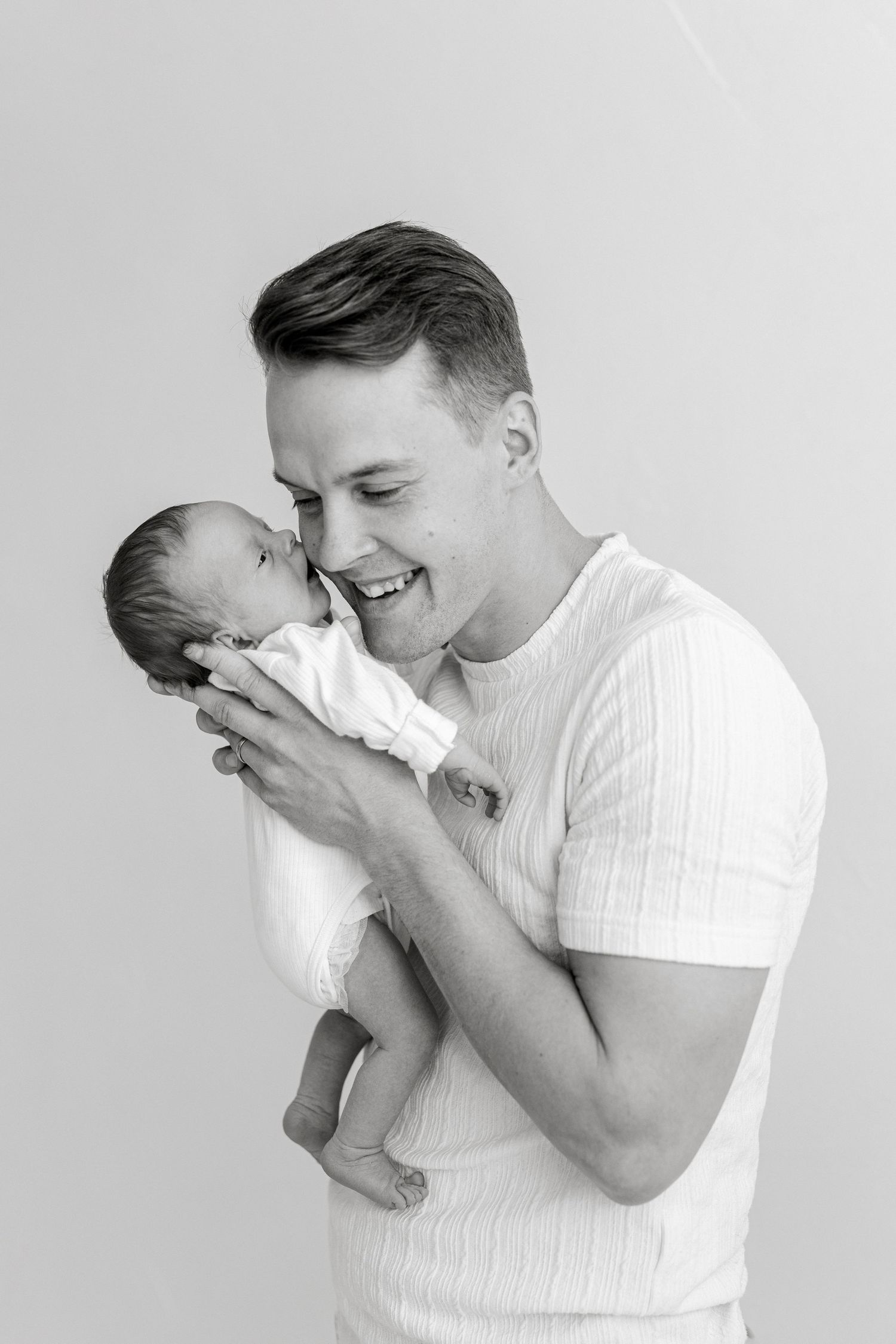 A smiling man in a white shirt tenderly holds and nuzzles a newborn baby in a black and white portrait.