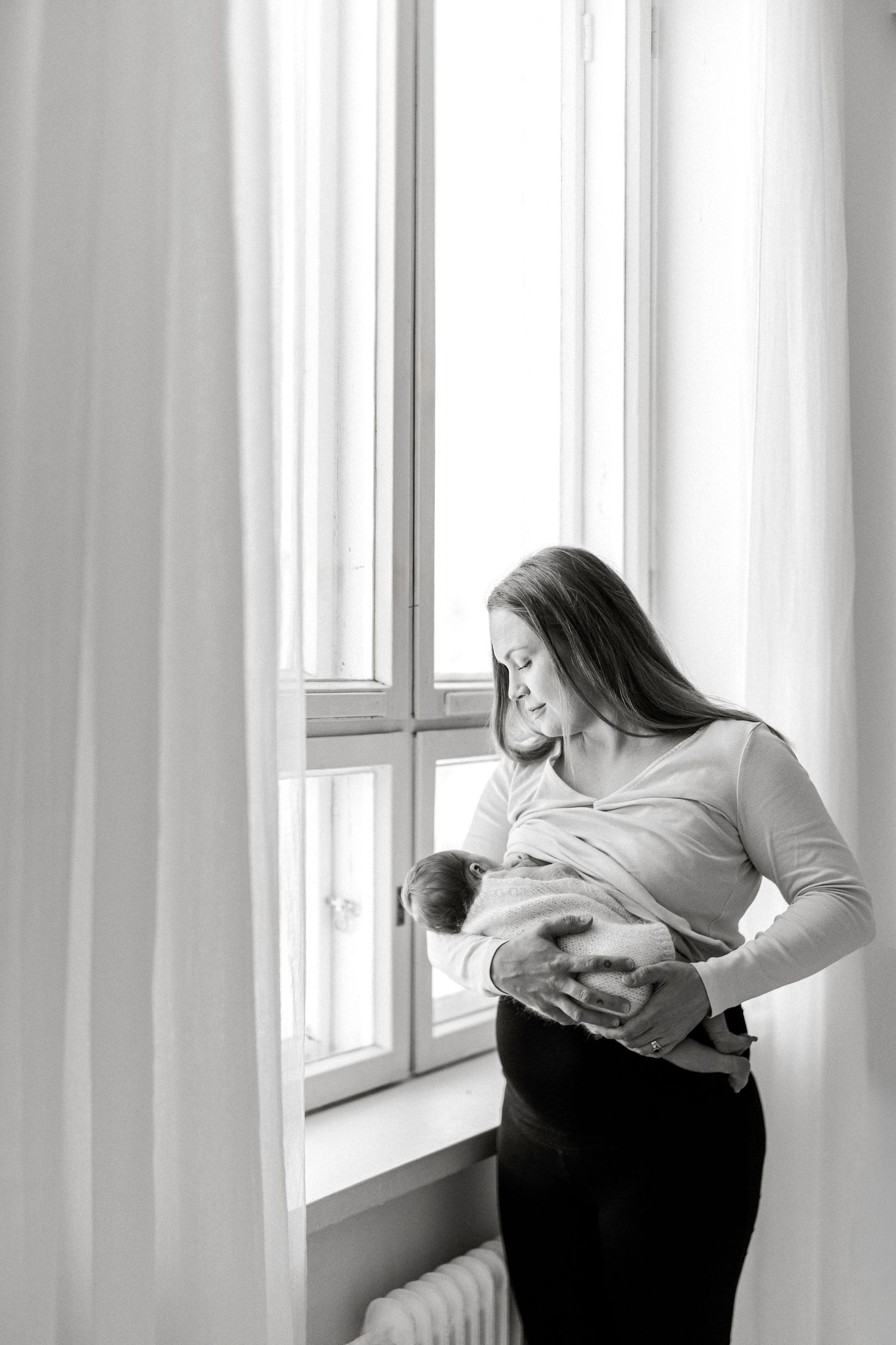 Black and white photo of a mother tenderly holding her newborn baby by a bright window.