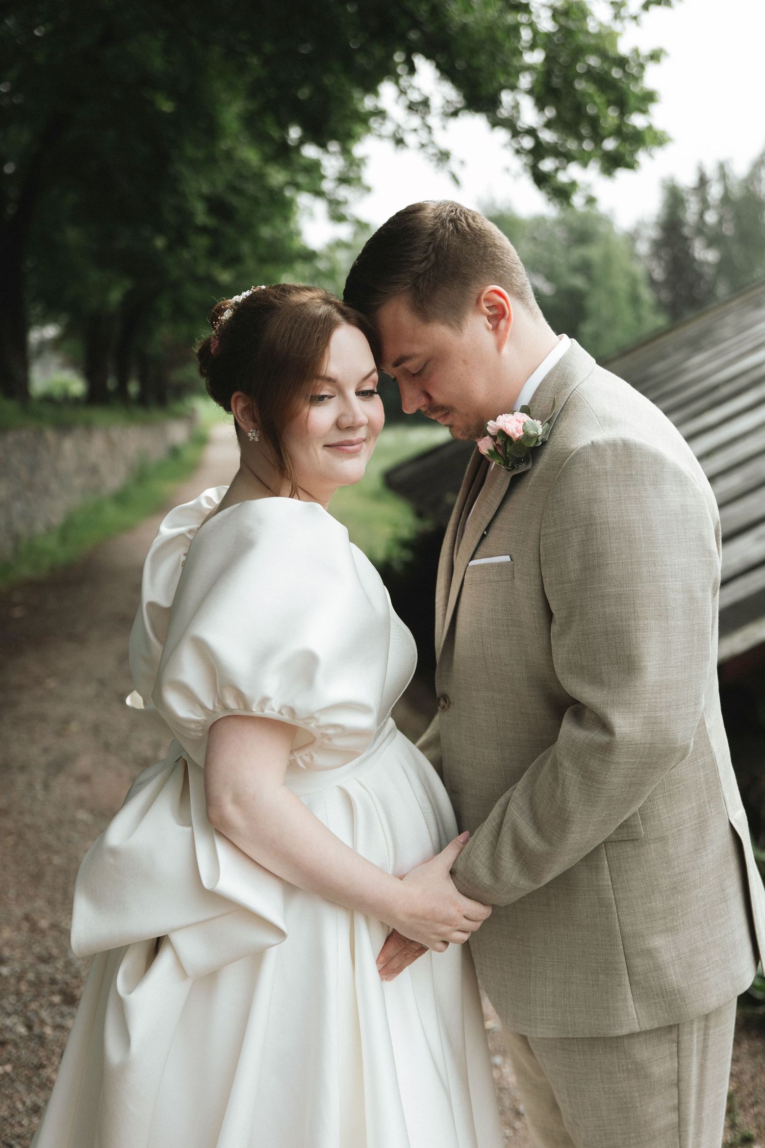 A bride in a white gown and groom in a beige suit share a tender moment outdoors, surrounded by lush green trees.