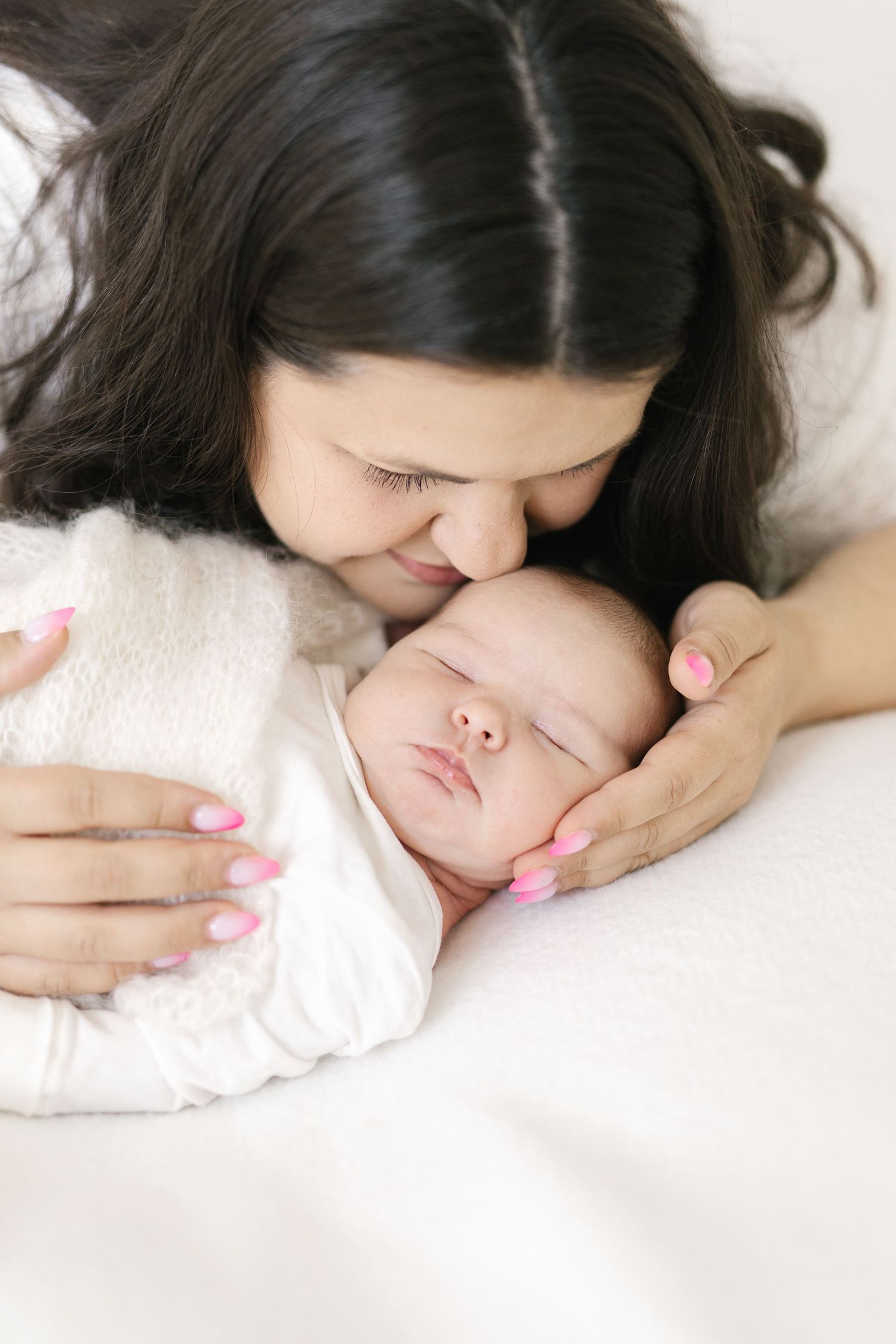 A loving mother with dark hair and pink nails gently kisses her sleeping newborn baby wrapped in a white blanket.