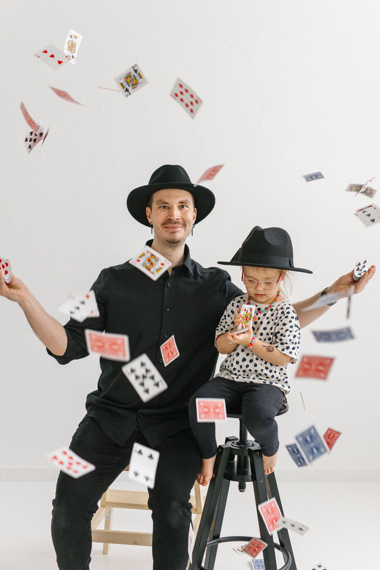 Magician and child performing card trick with playing cards floating in air against white background.