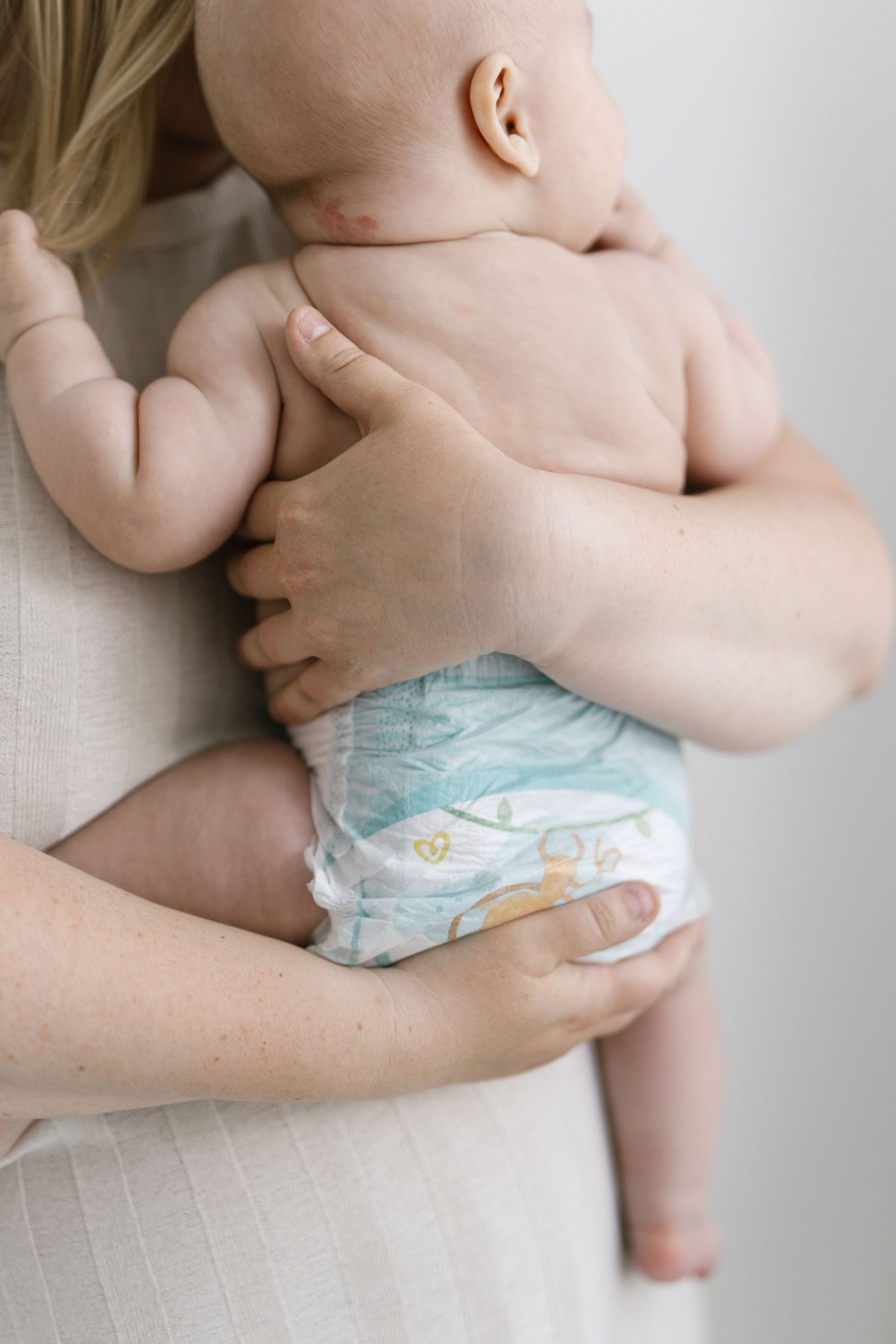 Mother holding a newborn baby in a diaper against her chest, showing the infant's bare back and chubby arms.