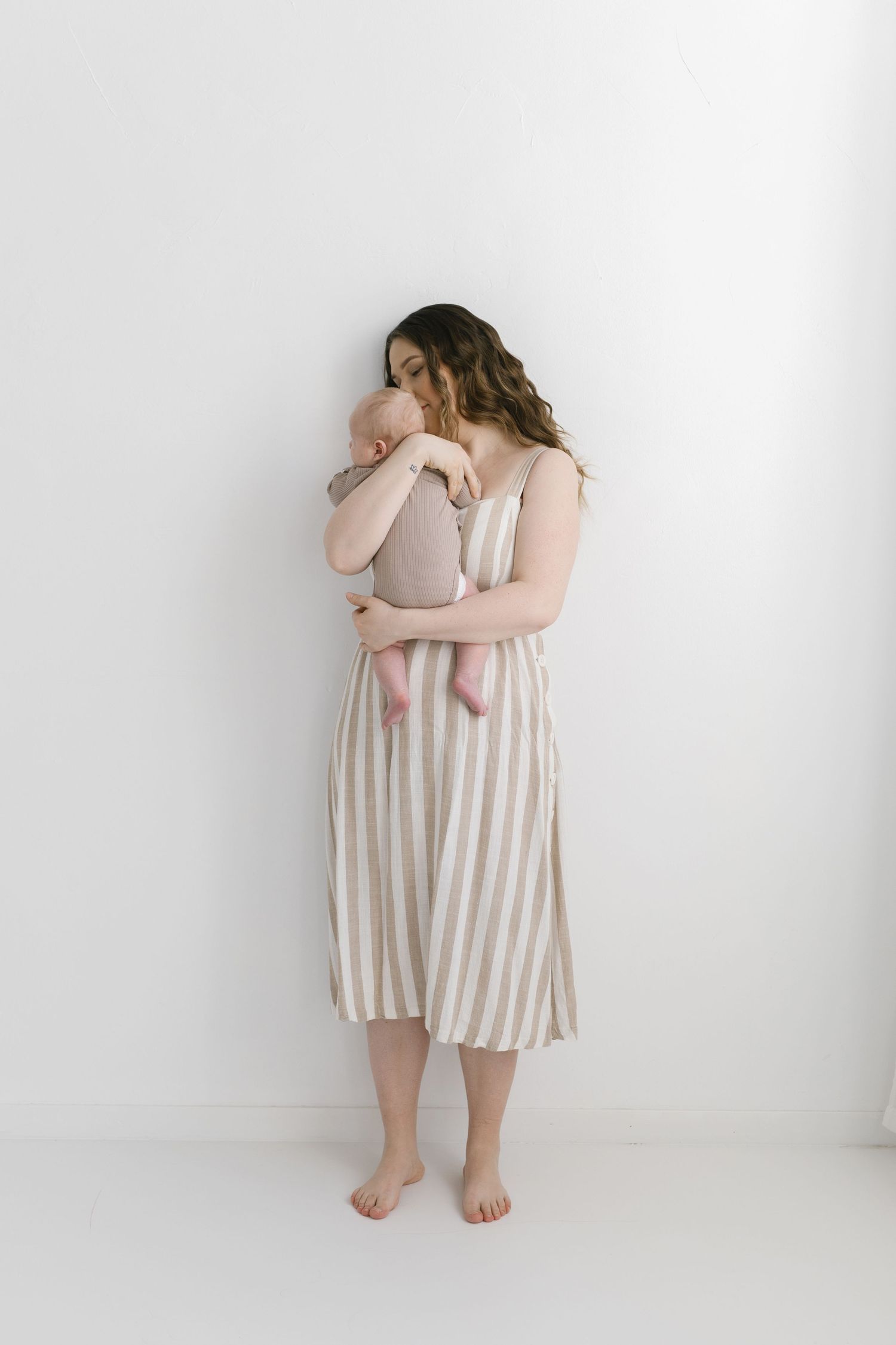 A barefoot woman in a striped midi dress tenderly holds a newborn baby against a white background.