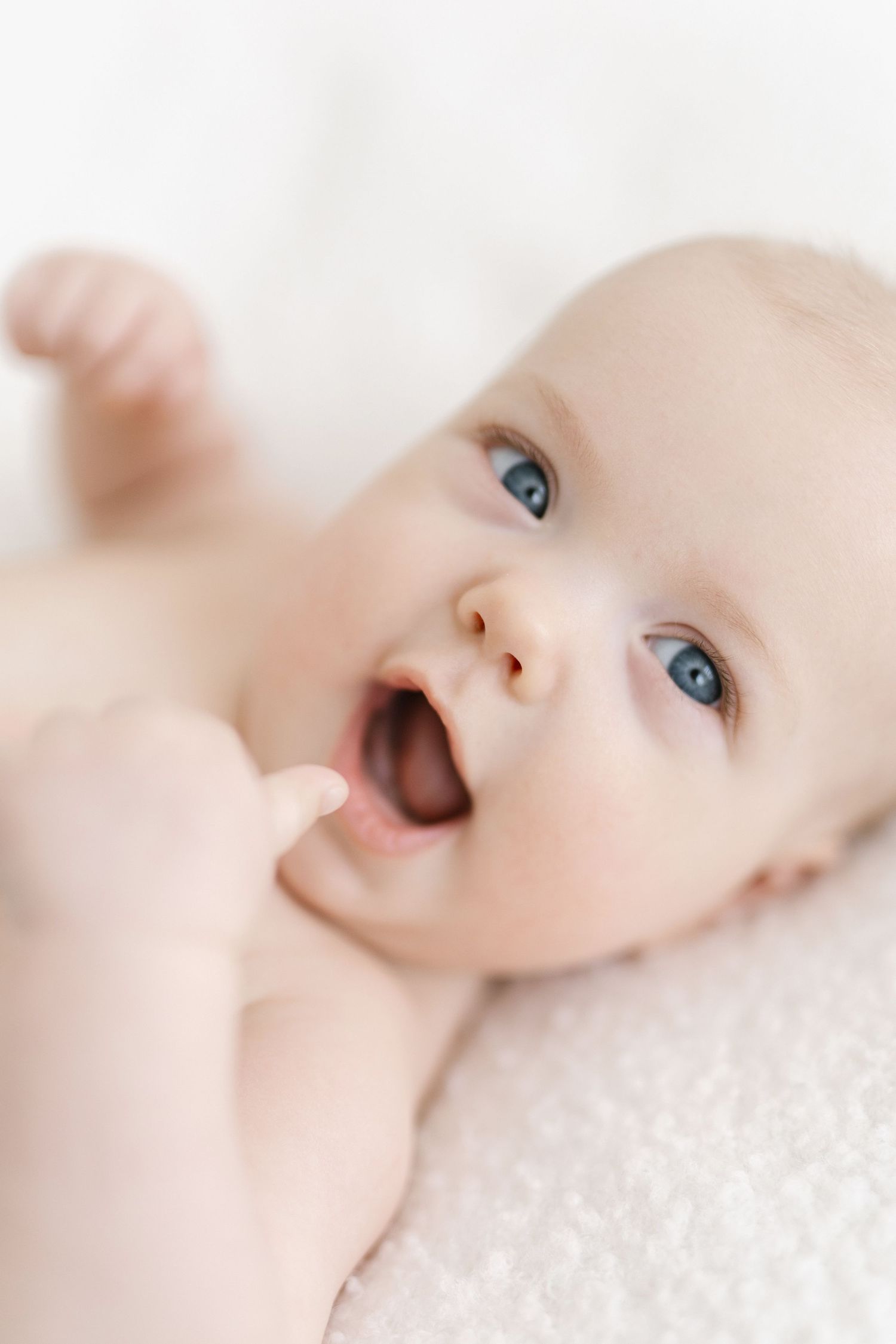 Adorable blue-eyed baby lying on white blanket with mouth open and hand raised, close-up portrait photo.