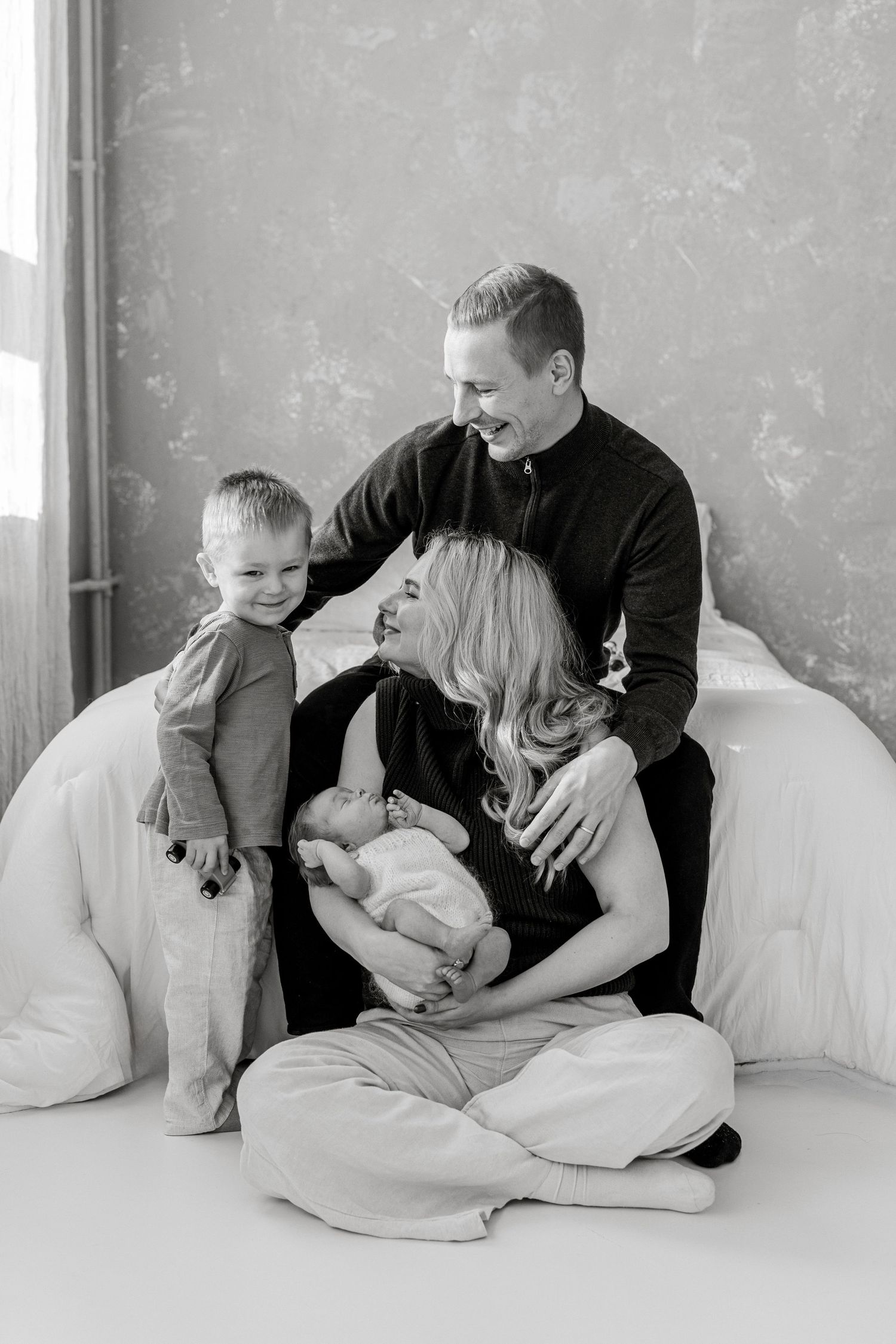 Black and white family portrait of smiling parents with a toddler boy and newborn baby posed together on a white bed in a studio.