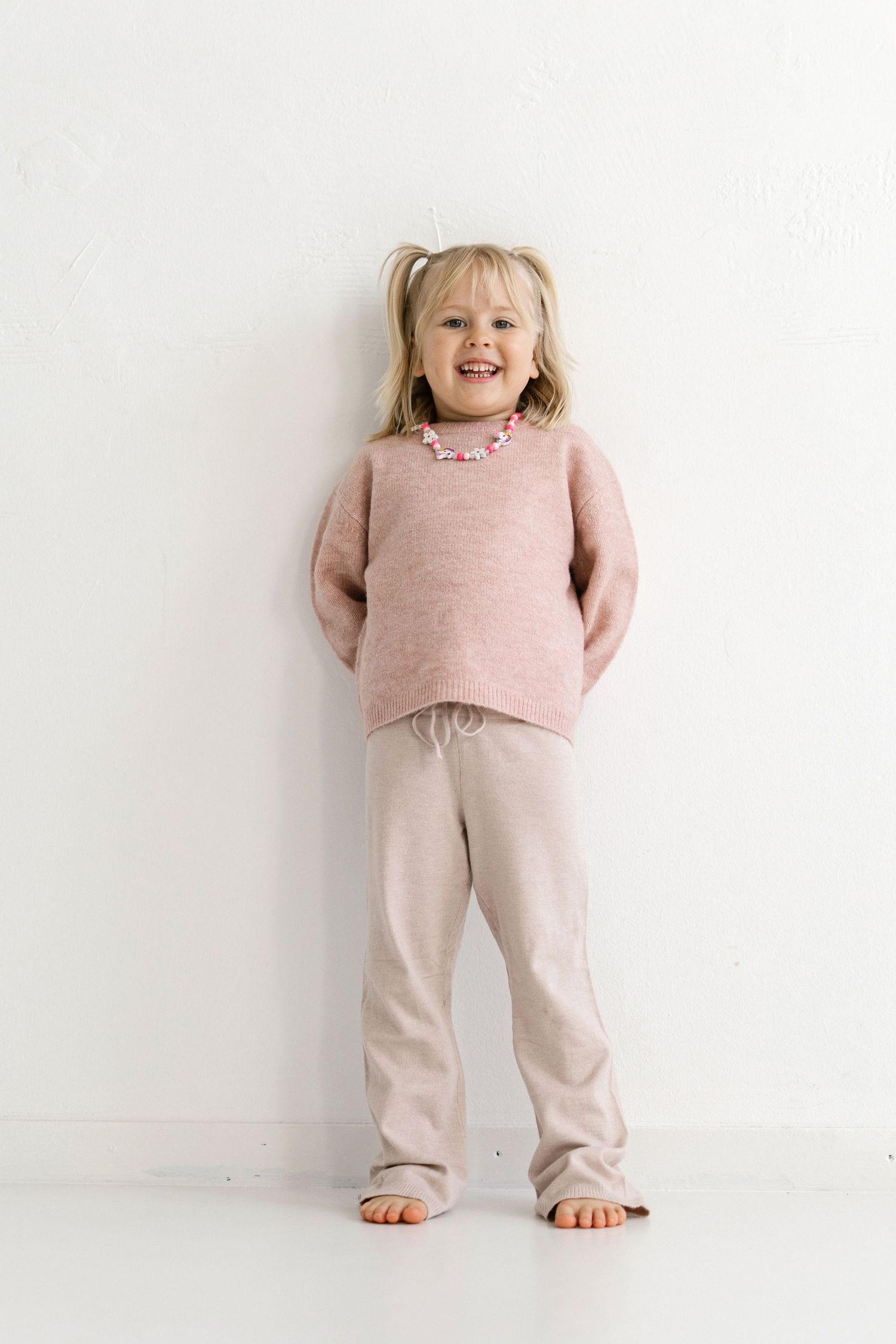 Smiling young girl in pink knit sweater and beige pants with pigtails, standing barefoot against white wall.