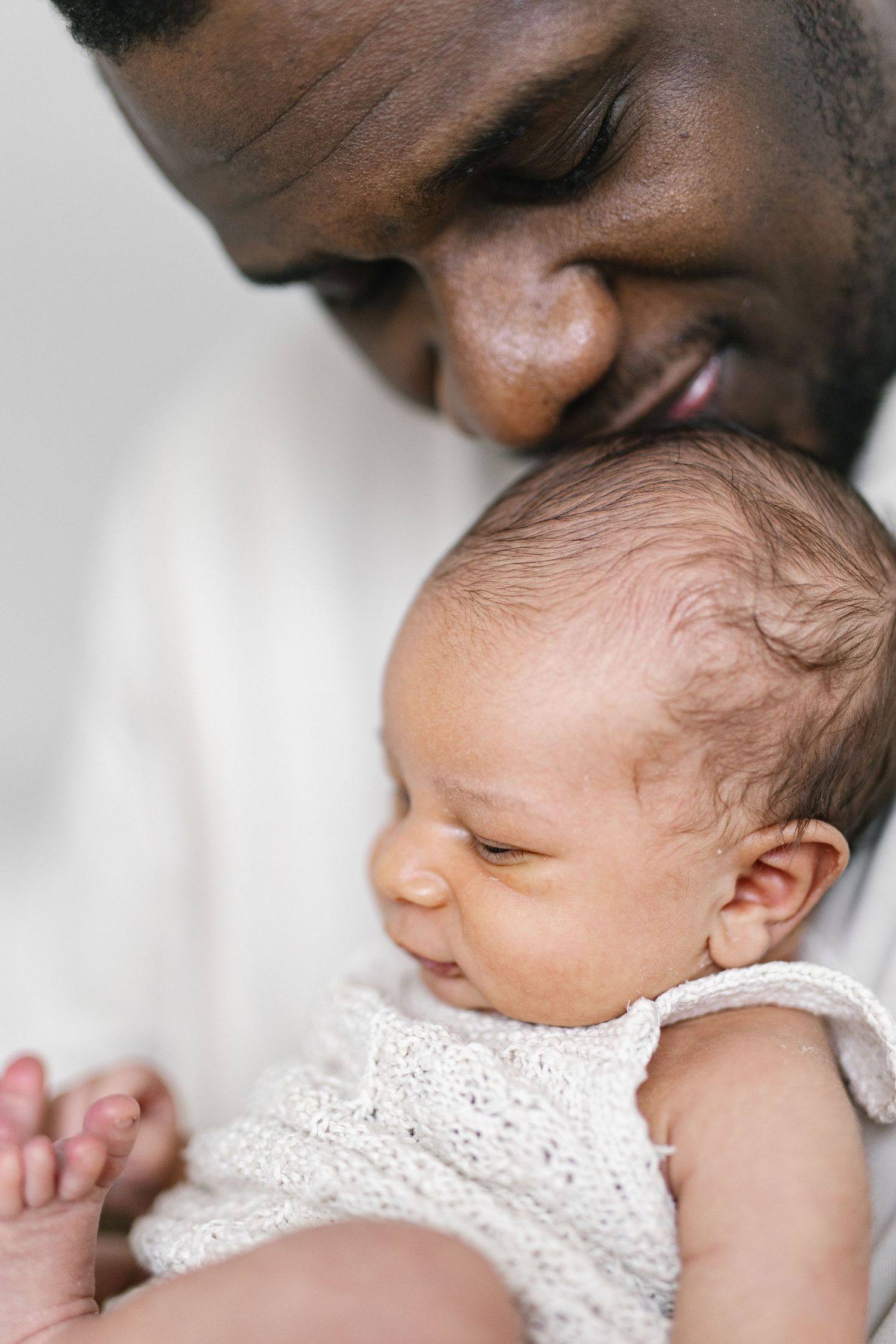 Close-up of tender moment between parent and sleeping infant in white dress.