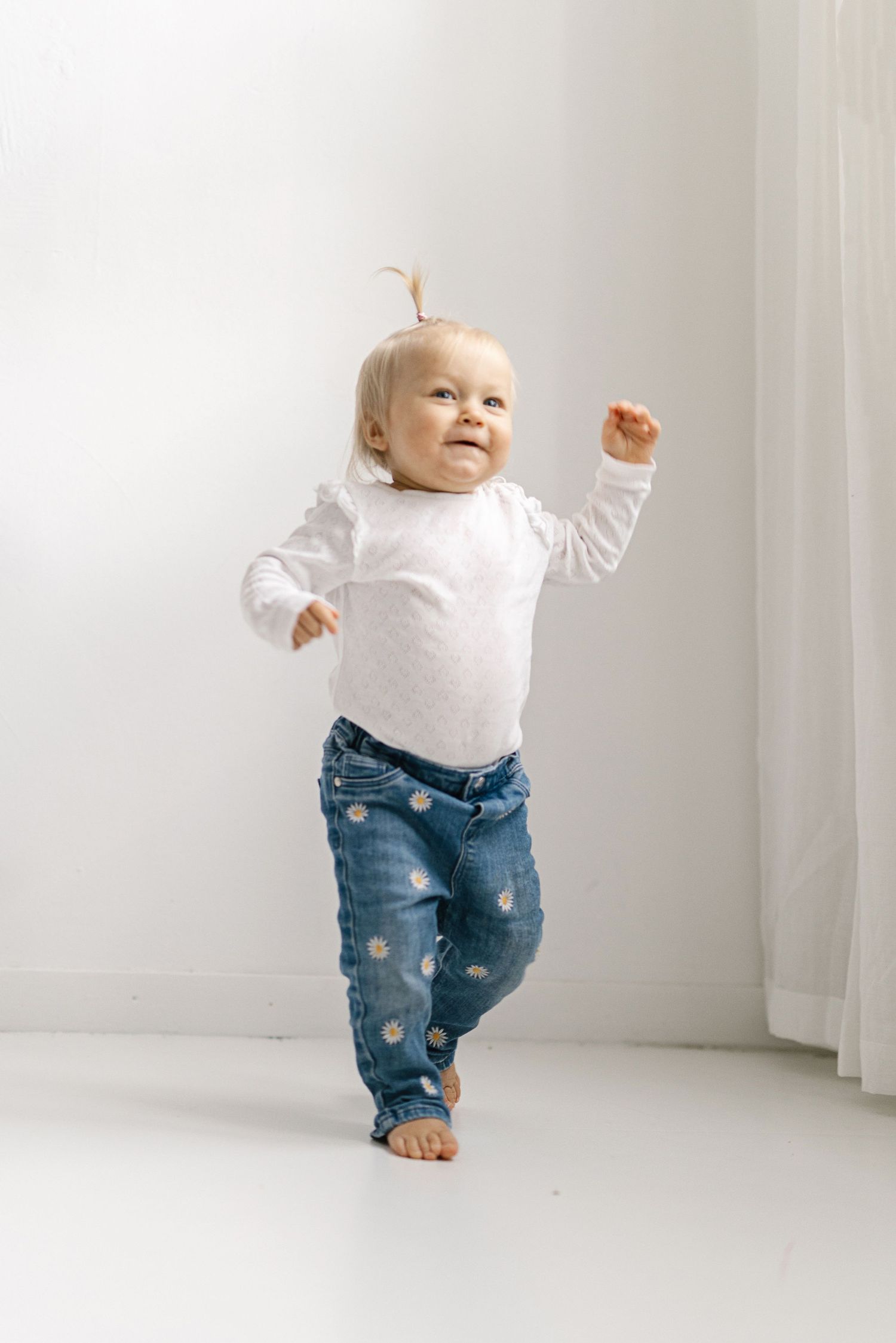 Adorable baby girl in white top and polka dot jeans taking first steps on white floor.