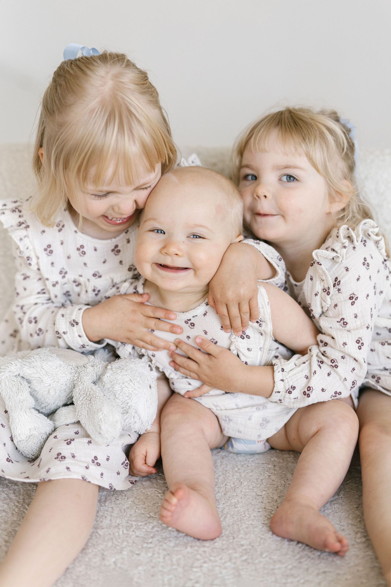 Three adorable blonde toddler girls in matching floral dresses cuddle together on a soft blanket, smiling joyfully.