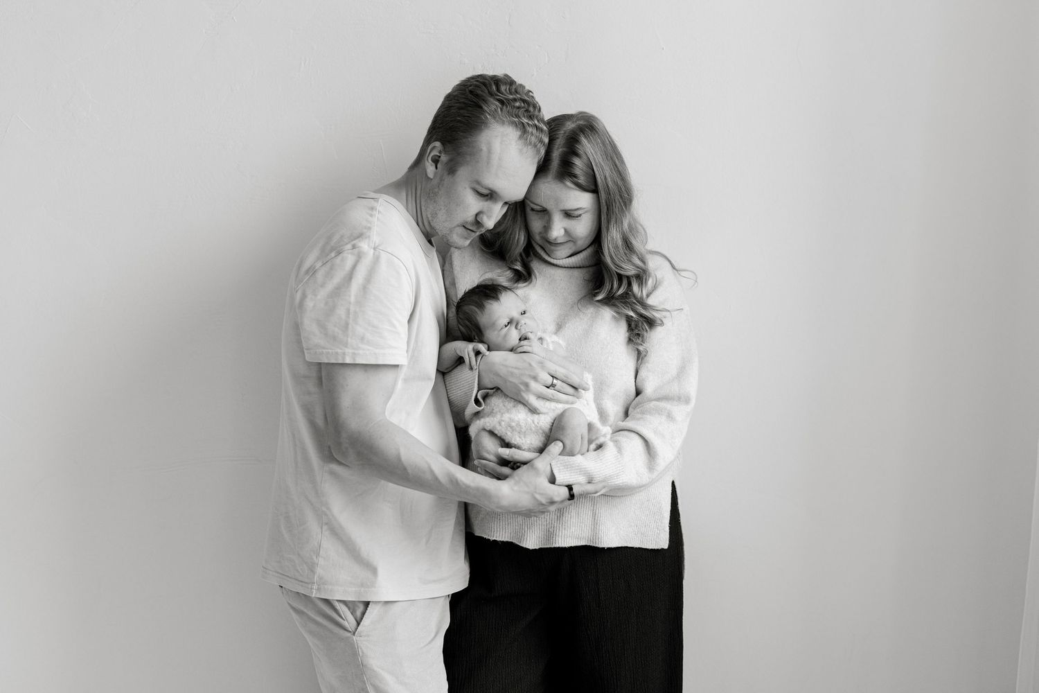 Black and white newborn portrait of new parents embracing and gazing down at their sleeping baby against a plain studio wall.