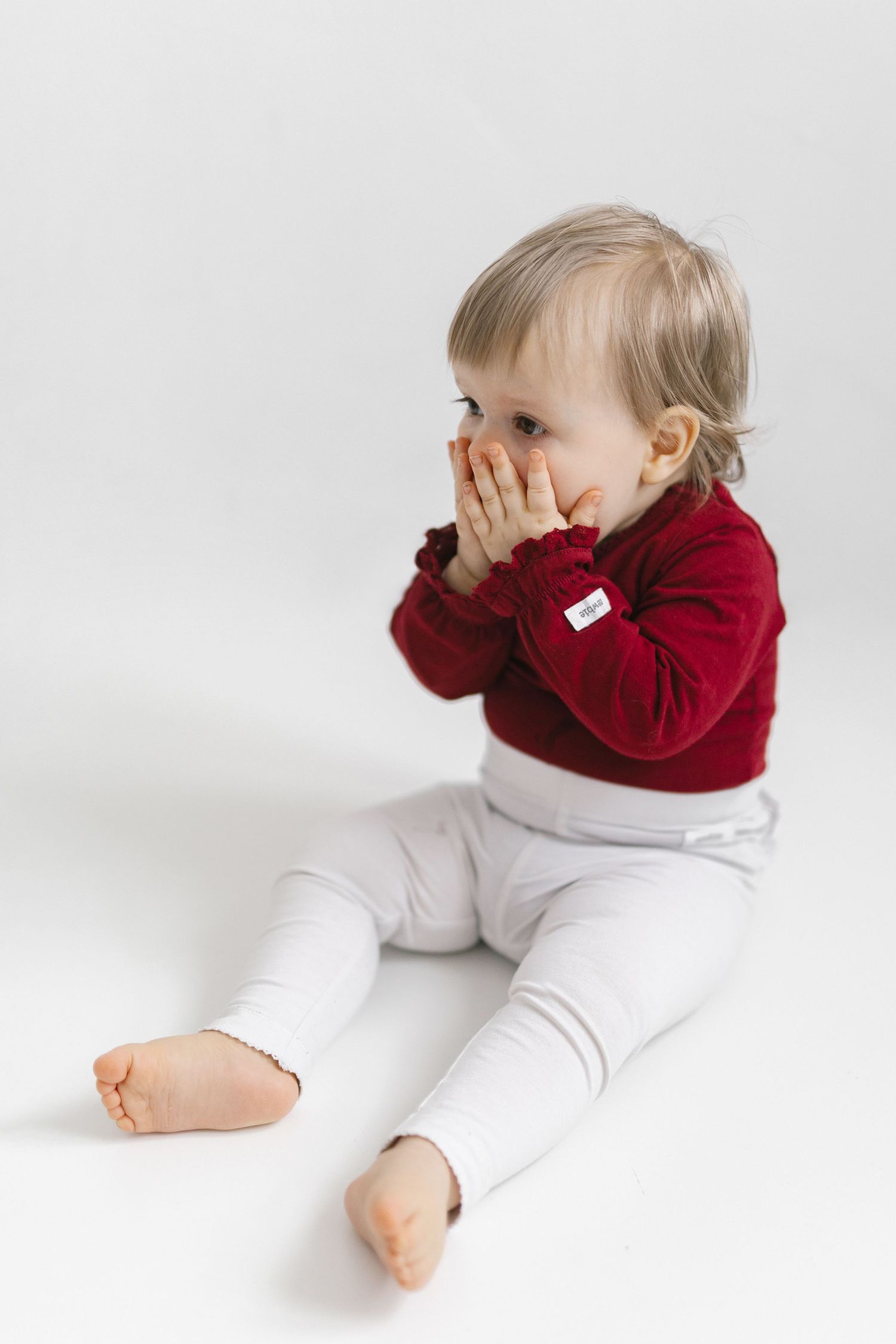 Baby in red velvet top and white pants plays peek-a-boo against white background in sequential photos.