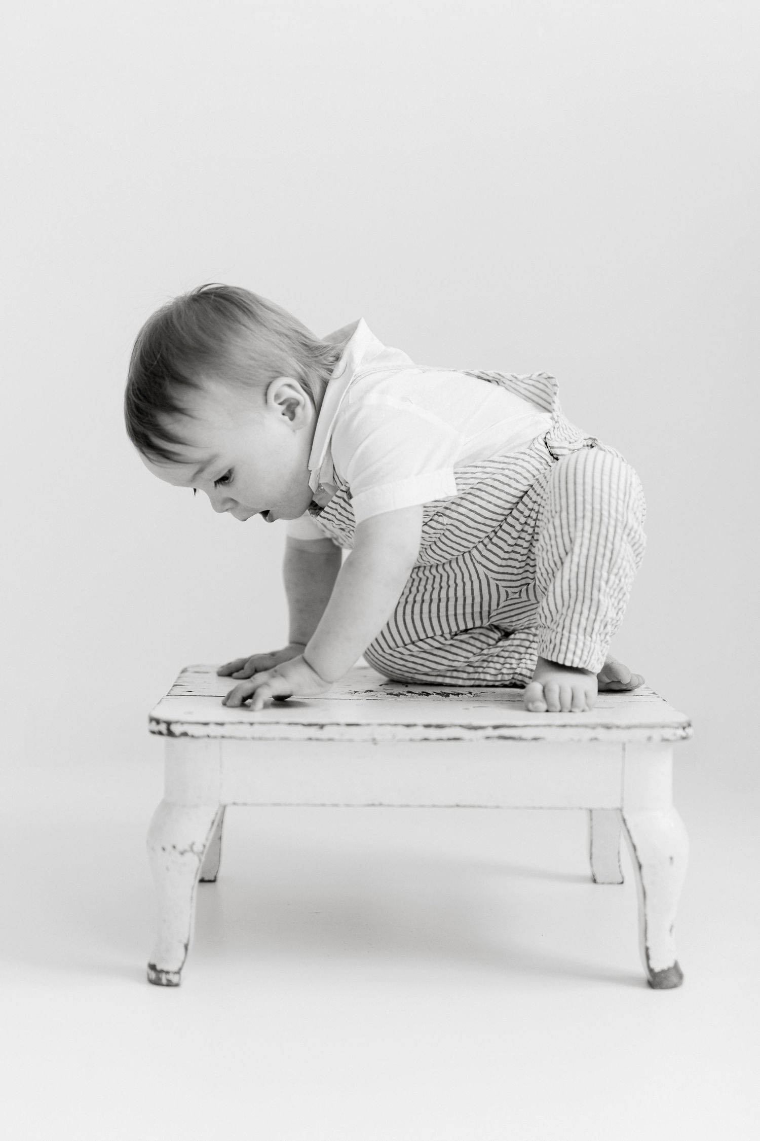 Baby in striped outfit crawling on white bench in studio setting.