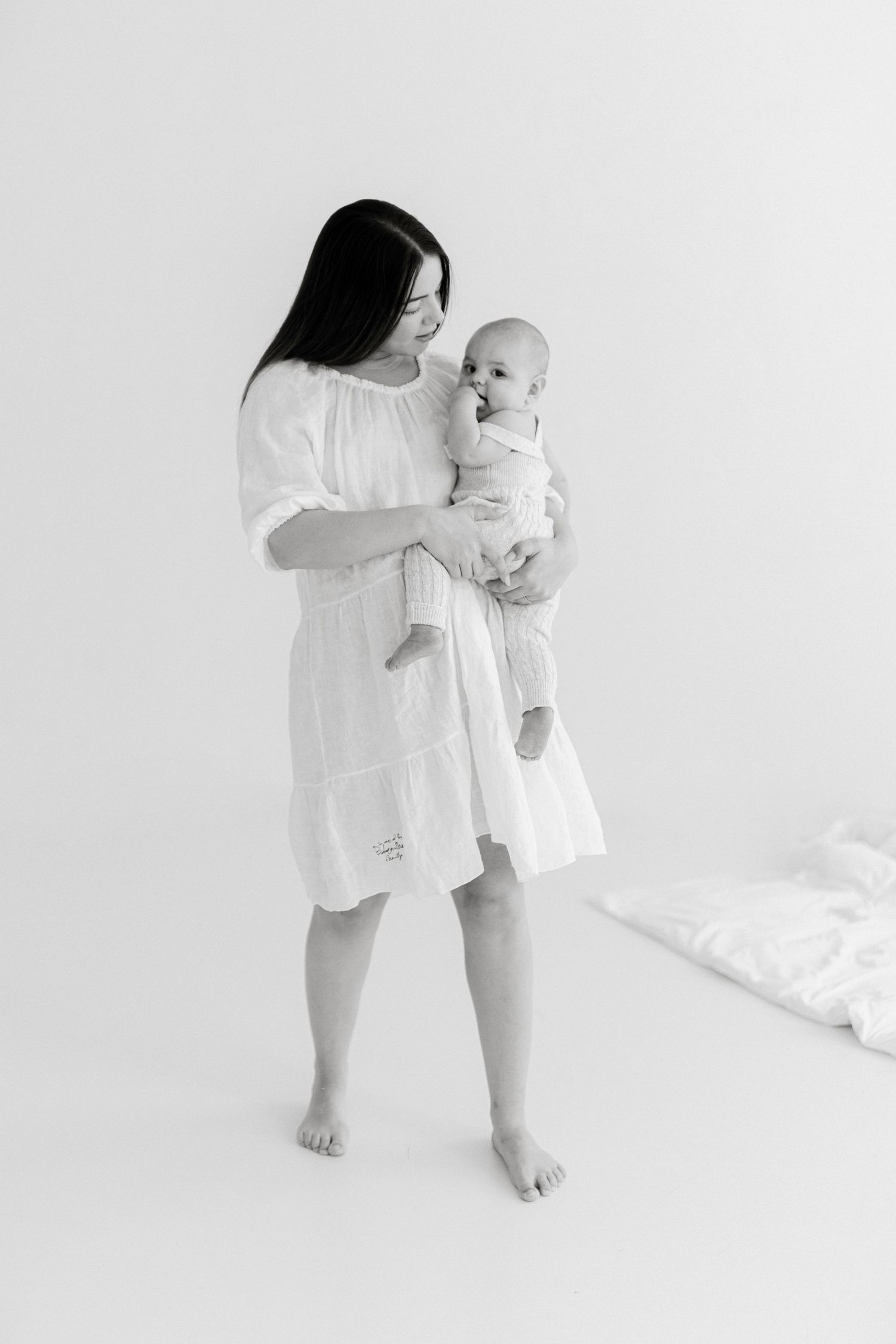 Black and white photo of a mother in a white dress holding her baby in a bright studio setting.