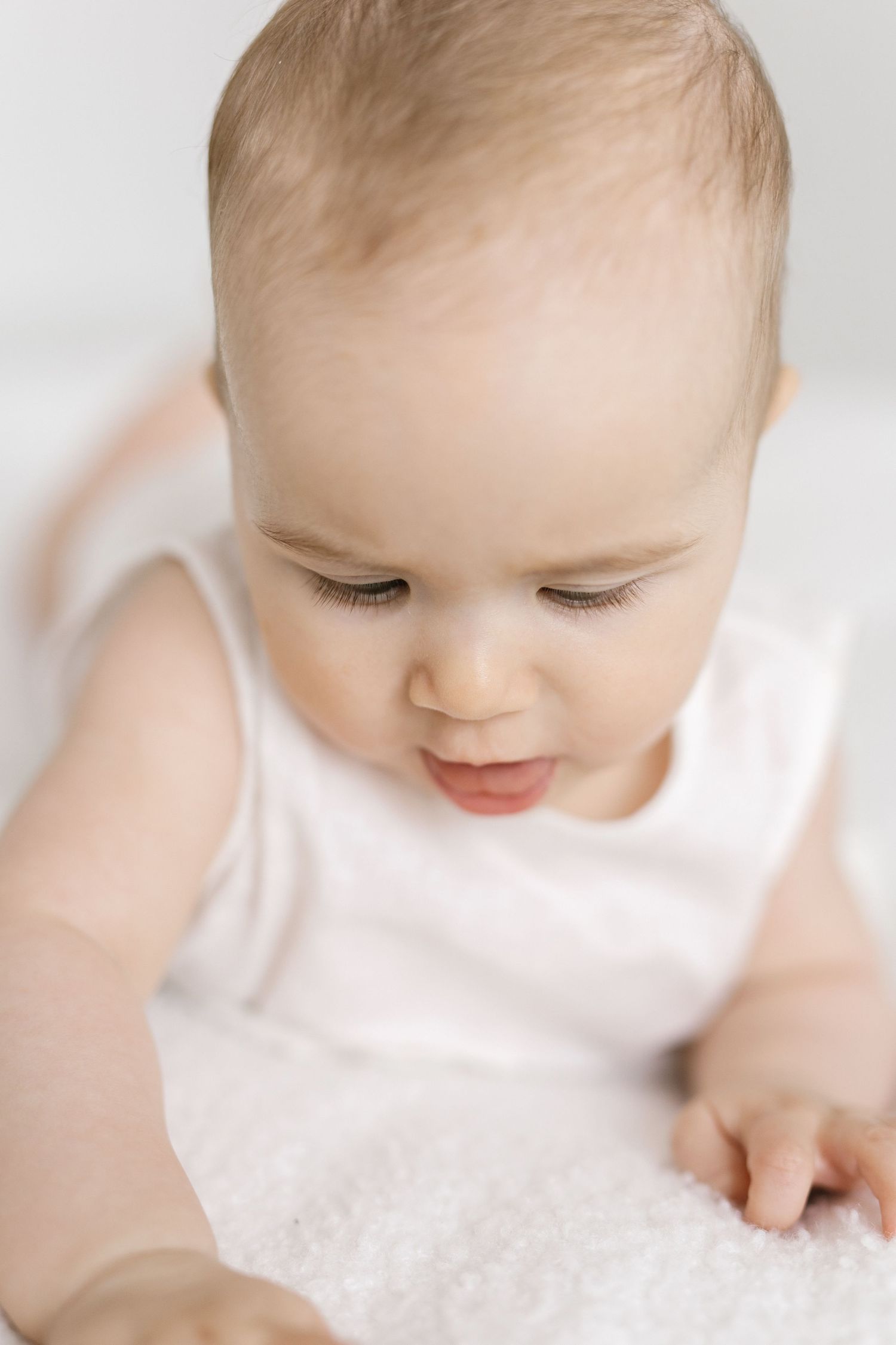 Adorable baby in white onesie looking down while lying on a soft white surface.
