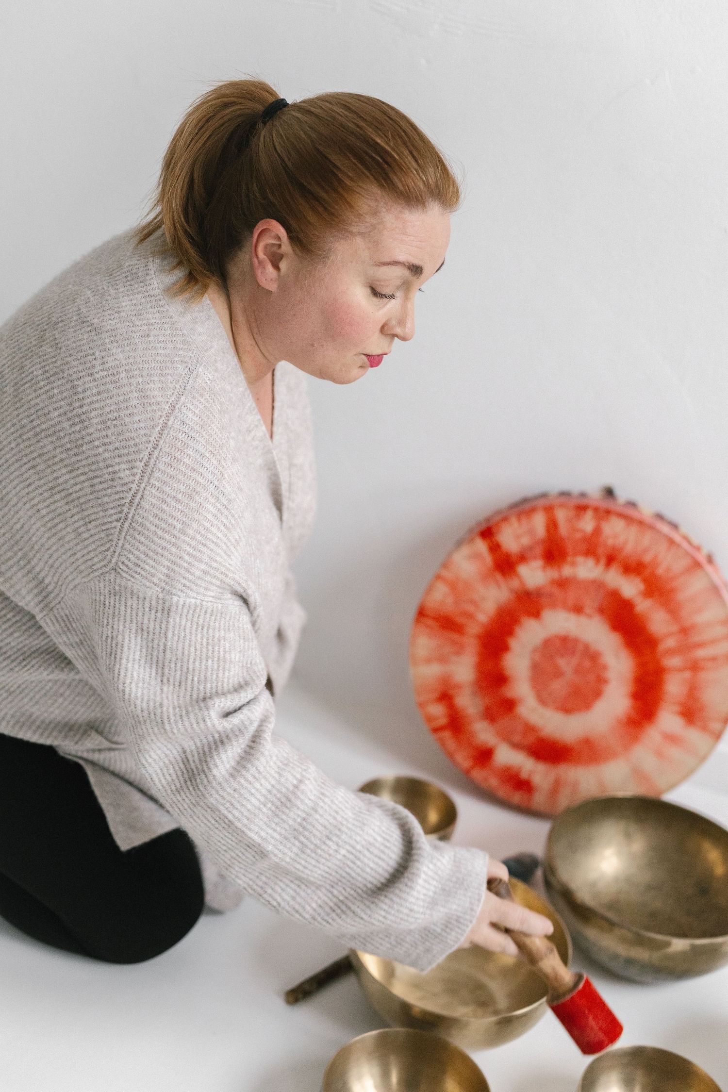 A person in a beige sweater plays Tibetan singing bowls while holding a red meditation cushion against a white wall.